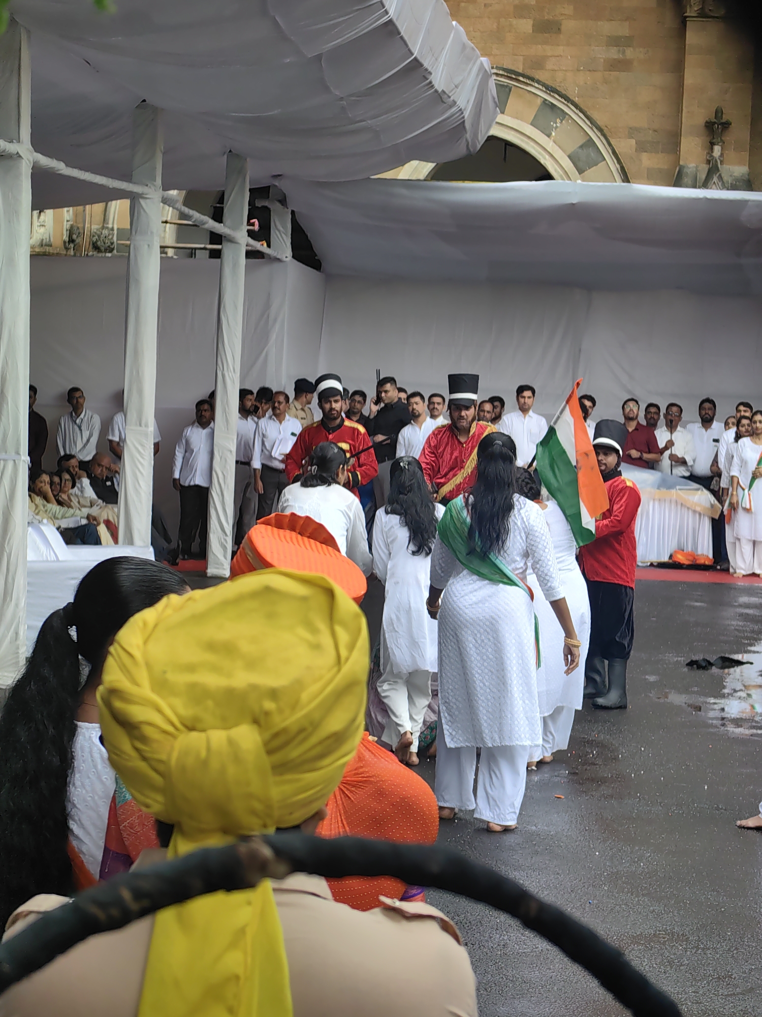 The image depicts an outdoor event in front of a brick building with arched windows, likely a historical structure in Mumbai, India. A white canopy covers part of the scene, supported by white pillars. In the foreground, a person wearing a yellow turban is partially visible, holding a dark object. Behind them, several individuals are dressed in white traditional Indian clothing. A group of performers, including two individuals in red jackets and black hats, are central to the image. One of them holds an Indian flag. The ground appears wet, suggesting recent rain. A crowd of onlookers is visible in the background, some seated and others standing. The overall scene suggests a ceremonial or cultural event.
