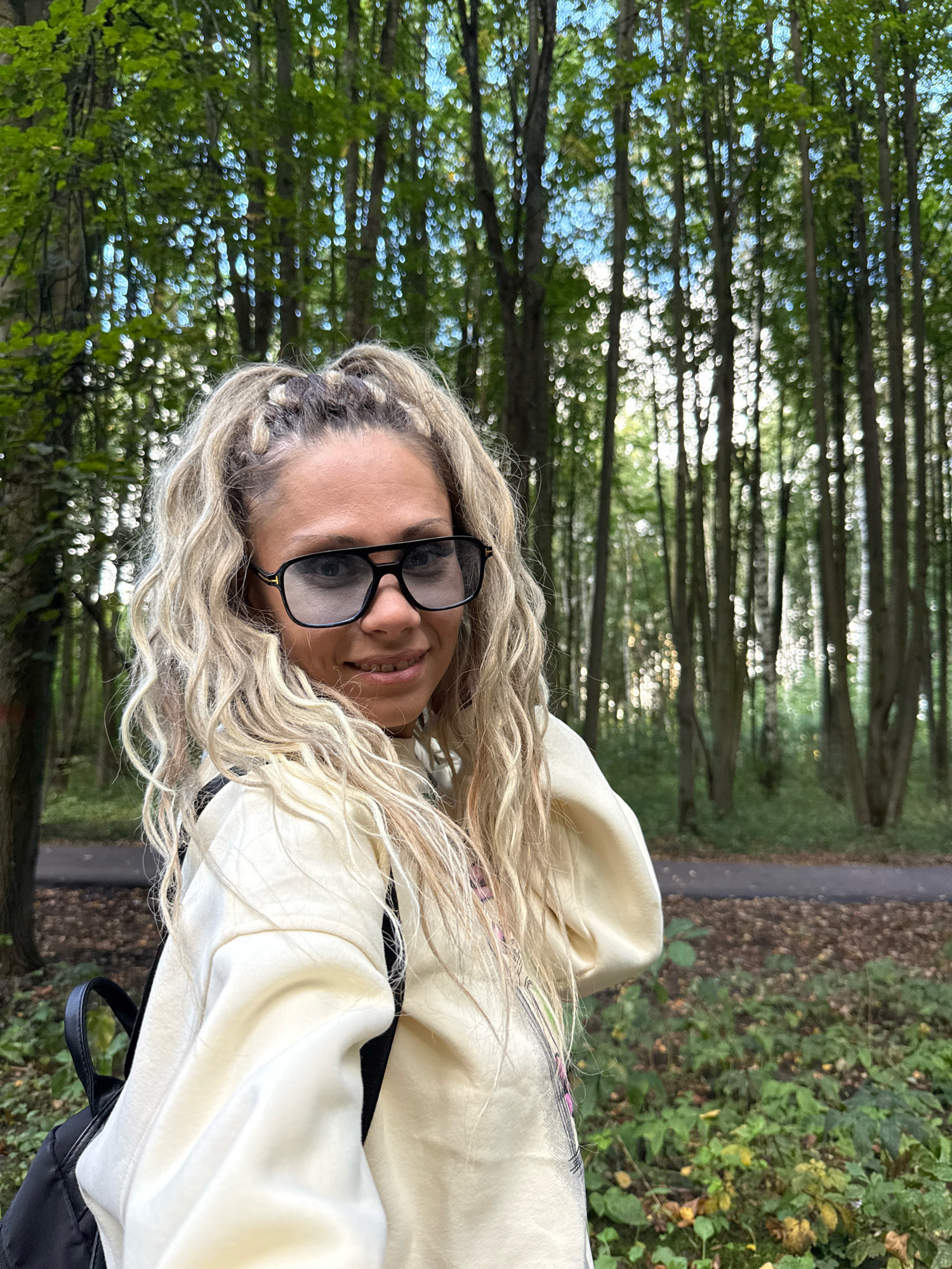 A woman with long blonde hair styled in braids and curls, wearing oversized black-rimmed sunglasses and a light yellow hooded sweatshirt, stands in a forest. The background features a dense arrangement of tall trees with green foliage, suggesting a park or wooded area. The ground is covered with fallen leaves and green undergrowth. A path can be seen in the distance. The woman is looking at the camera and smiling. A black backpack is visible over her shoulder. The lighting suggests it is daytime. The location is identified as Rusanovo, Russia.