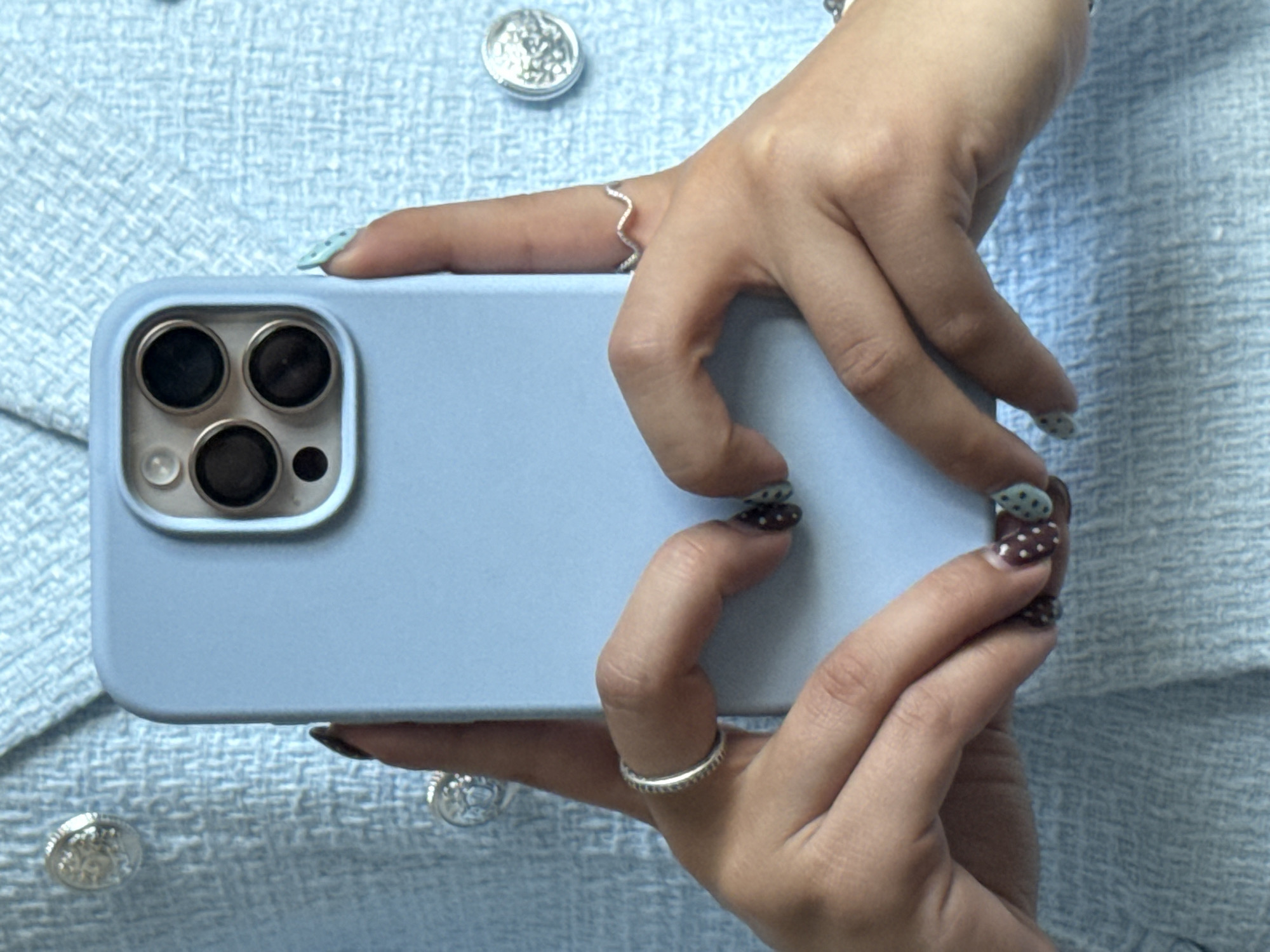 A person is holding a light blue smartphone with a visible camera module featuring three large lenses and a smaller sensor or flash. The phone is held horizontally, with the camera facing left. The person's hands, adorned with rings and patterned nail polish, are gripping the edges of the phone. The person is wearing a light blue tweed-like jacket or top with decorative silver buttons. The background is the same light blue tweed-like fabric. There is no geographical information about Bruchköbel, Germany in the image.