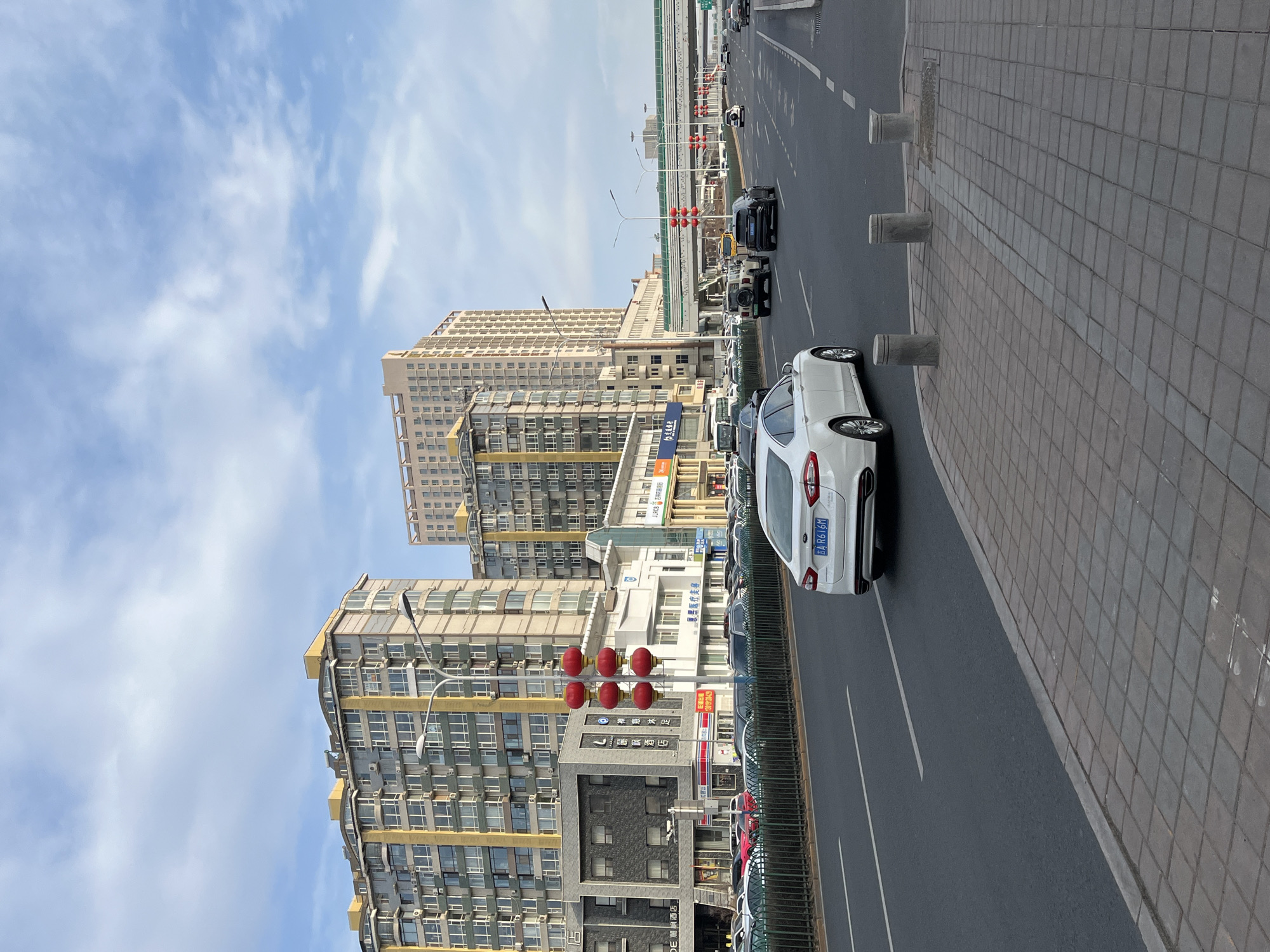 The image depicts a street scene in Changchun City, China. The photograph is taken at street level on the sidewalk, looking out to the street with buildings in the background. On the right of the image is a sidewalk paved with rectangular blocks of gray stone and a street with a white car in the foreground. Several other cars are further down the street. A concrete barrier separates the sidewalk from the street. Tall buildings are visible on the left side of the image, including residential apartment buildings and commercial buildings. The sky is blue with light clouds. A set of four red lanterns is strung on a street light pole.