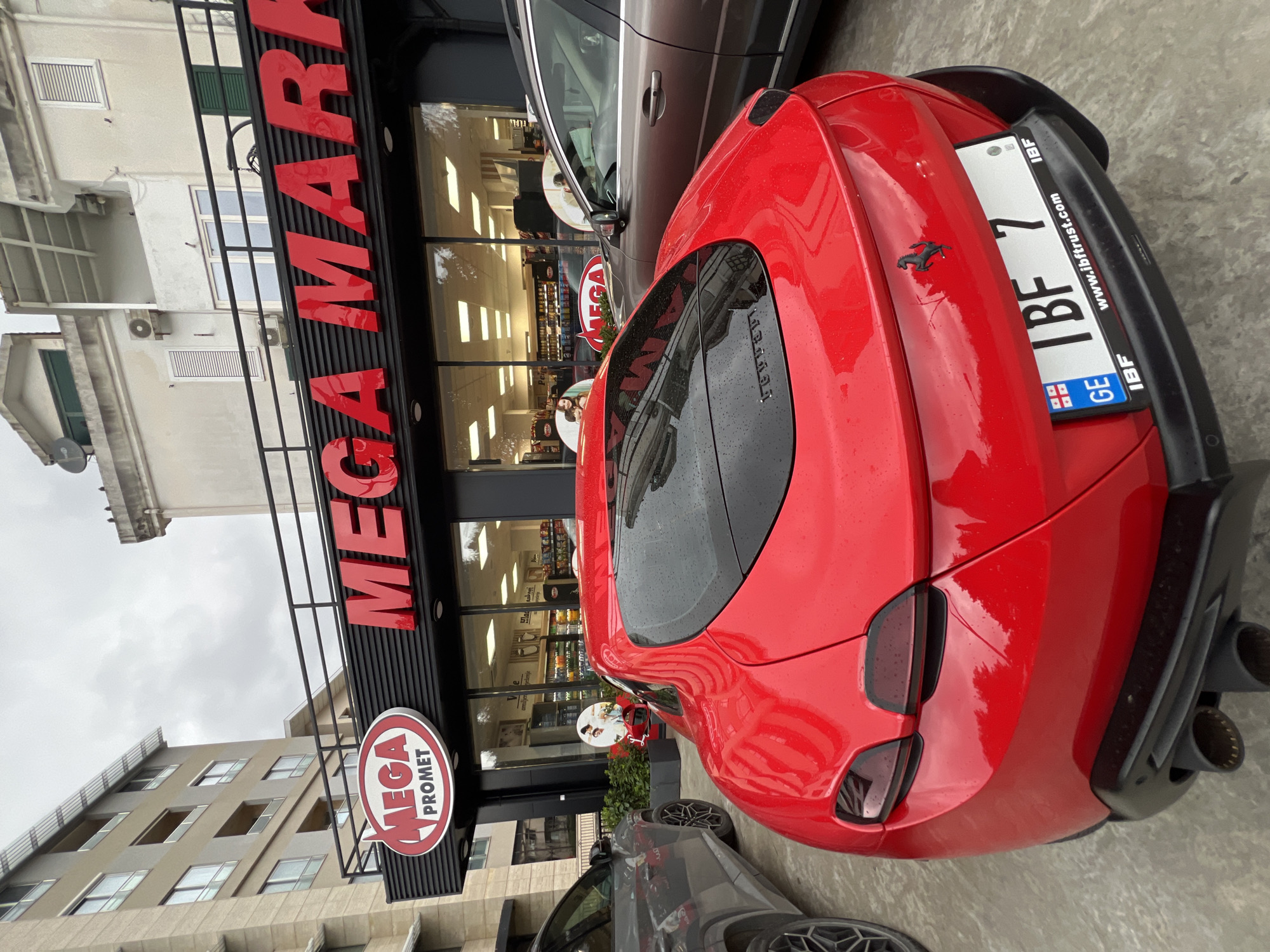 A red Ferrari Roma coupe, exhibiting water droplets on its body, is parked on a paved surface. The vehicle's rear faces the viewer, displaying a Georgian license plate "GE IBF 7" with "IBF" and "www.ibftrust.com" on the plate frame, and a black Ferrari prancing horse emblem above it. To the left, a grey SUV is partially visible, and to the right, a brown SUV is partially visible. The Ferrari is situated directly in front of a "MEGA MARKET" supermarket, identified by large red illuminated signage. A smaller "MEGA PROMET" sign is also visible above the store's entrance. The supermarket features large glass storefront windows reflecting internal lighting and product displays. Decorative circular signs are present near the entrance. Behind the supermarket, an older multi-story building with windows and an external air conditioning unit is visible, while a modern multi-story building with balconies stands to the left. The sky is overcast. This scene is located in Budva, Montenegro, in front of a Mega Market grocery store.