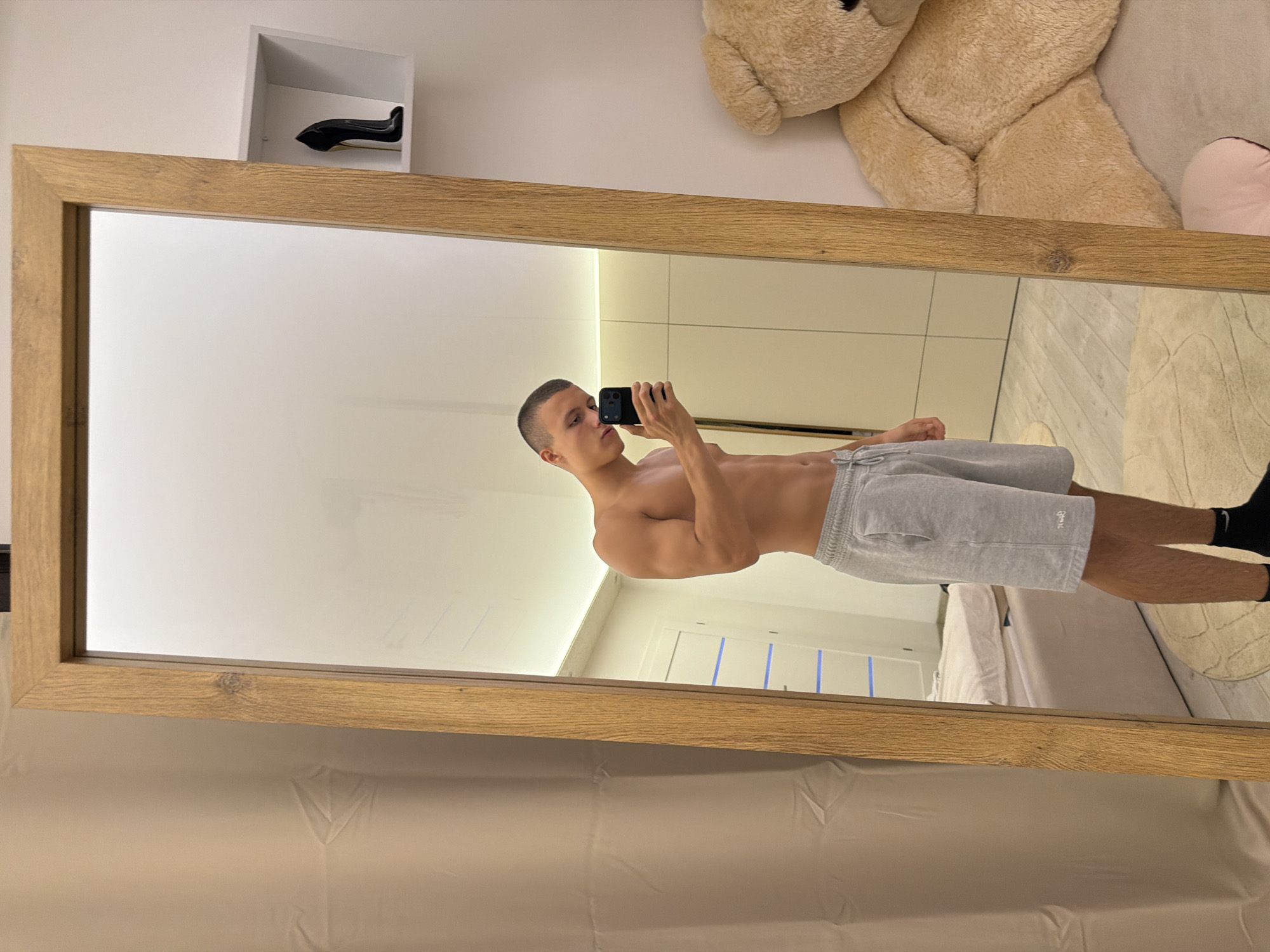 A young man with a short haircut is taking a mirror selfie in a room. He is shirtless, wearing grey athletic shorts and black socks. His left arm is bent, holding a smartphone to capture the reflection. To the right of the mirror, a large, light brown teddy bear is visible, along with a shelf on the wall holding a black high-heeled shoe. The room appears to be a bedroom, with a bed and a closet visible in the reflection. The floor is covered with a rug. The scene is set in an apartment in Elhovo, Bulgaria.