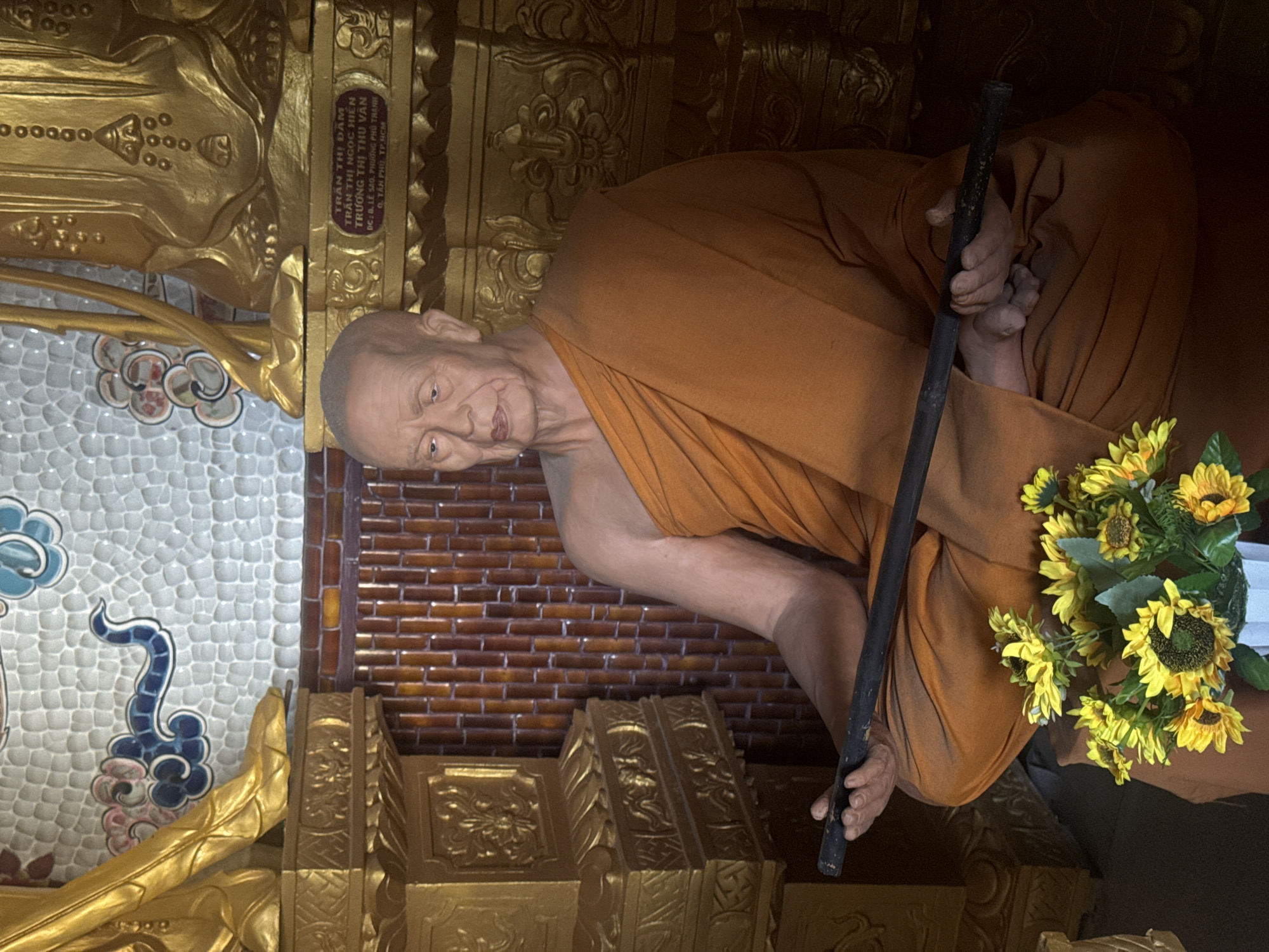 This image depicts a life-sized statue of an elderly Buddhist monk, seated cross-legged on a dark platform in what appears to be a temple or shrine, located in the city of Xuân Thành, Vietnam.
The monk statue, rendered with realistic detail, has a bald head and a weathered face featuring pronounced wrinkles on the forehead, around the eyes, and mouth, consistent with advanced age. His eyes are open, gazing slightly downward and to the viewer's left, with a serene or contemplative expression. He is draped in a loose-fitting, saffron or burnt orange monastic robe (kasaya) that covers his body from the shoulders down, exhibiting natural folds and creases. Both hands are positioned on his lap, holding a dark, cylindrical staff or rod horizontally across his knees.
In the immediate foreground, at the bottom center, a white vase containing a bouquet of artificial sunflowers is placed. The sunflowers are bright yellow with dark brown centers and green leaves.
The background features ornate architectural elements. To the left and right of the monk statue, large golden pedestals or bases are visible, intricately carved with decorative patterns. The lower sections of two standing golden statues are visible above these pedestals; the one on the left shows a distinct foot, while the one on the right shows a section of draped garment. On the golden base to the right, a rectangular plaque is affixed, displaying Vietnamese text in black: "TRẦN THỊ ĐÀM," "TRƯƠNG THỊ NGỌC HIỀN," "TRƯƠNG THỊ THU VÂN," "DC: 8, LÊ SAO, PHƯỜNG PHÚ THẠNH," and "Q. TÂN PHÚ, TP. HCM."
Behind the golden structures, the wall is composed of two distinct sections. The lower section consists of reddish-brown, vertically aligned, cylindrical elements, resembling bamboo or narrow wood planks. The upper section is a light-colored tiled wall with a textured, possibly scaled pattern. This tiled wall is decorated with stylized cloud motifs in blue and pink, and a blue serpentine or bird-like design on the left, and fainter circular patterns on the right.