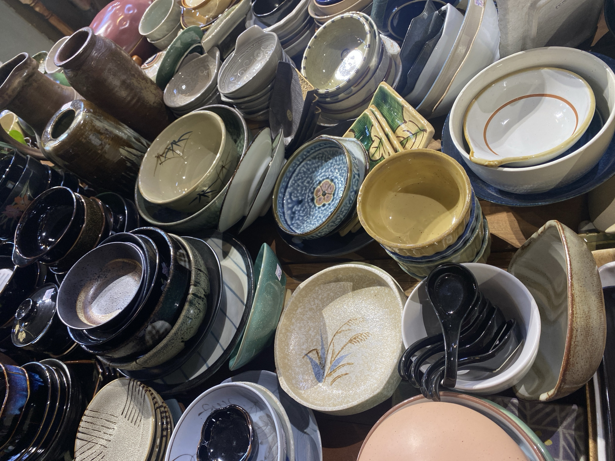 The image depicts a close-up, top-down view of a collection of ceramic dishes, bowls, and jars, densely packed and arranged in a layered fashion. The items are primarily in earthy tones of brown, beige, and grey, with some accents of blue and black. Many of the dishes feature traditional Japanese or East Asian patterns, such as geometric lines, floral motifs, and abstract designs. Several stacks of plates are visible, along with various sizes and shapes of bowls, some appearing to be serving bowls and others smaller, individual portions. A few larger ceramic jars, one with a wide opening and another taller and more slender, are prominent in the background. The lighting is somewhat dim, suggesting an indoor setting, possibly a market stall or a retail display. There is no indication of the specific location in Nha Trang Ward, Vietnam, beyond what can be inferred from the type of goods displayed, which are common in artisanal markets. No people are visible in the image.