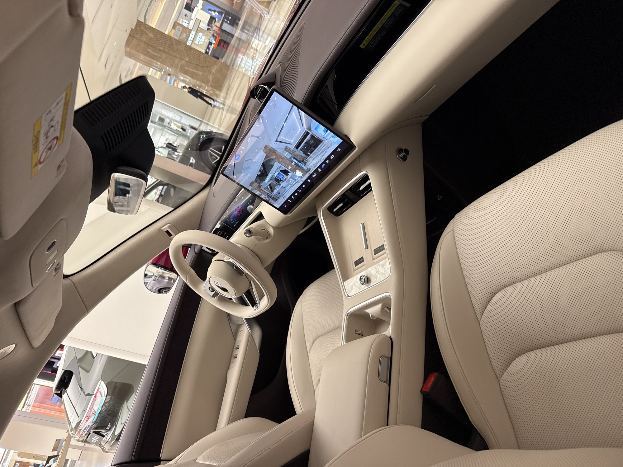 This is a photograph taken inside a luxury vehicle, likely a showroom, with the view directed towards the dashboard and passenger seats. The interior is dominated by cream-colored leather upholstery and brushed metal accents. A large, modern touchscreen display is prominently featured on the dashboard, showing a 360-degree view of the surrounding environment, which appears to be a brightly lit, upscale shopping mall. Other visible elements include the steering wheel, center console with gear selector, and the passenger seat. Through the car's windows, glimpses of other vehicles and shoppers in the mall can be seen. The overall impression is one of sophistication and advanced technology. There is no specific visual information in the image to confirm the location as Huizhou, China.