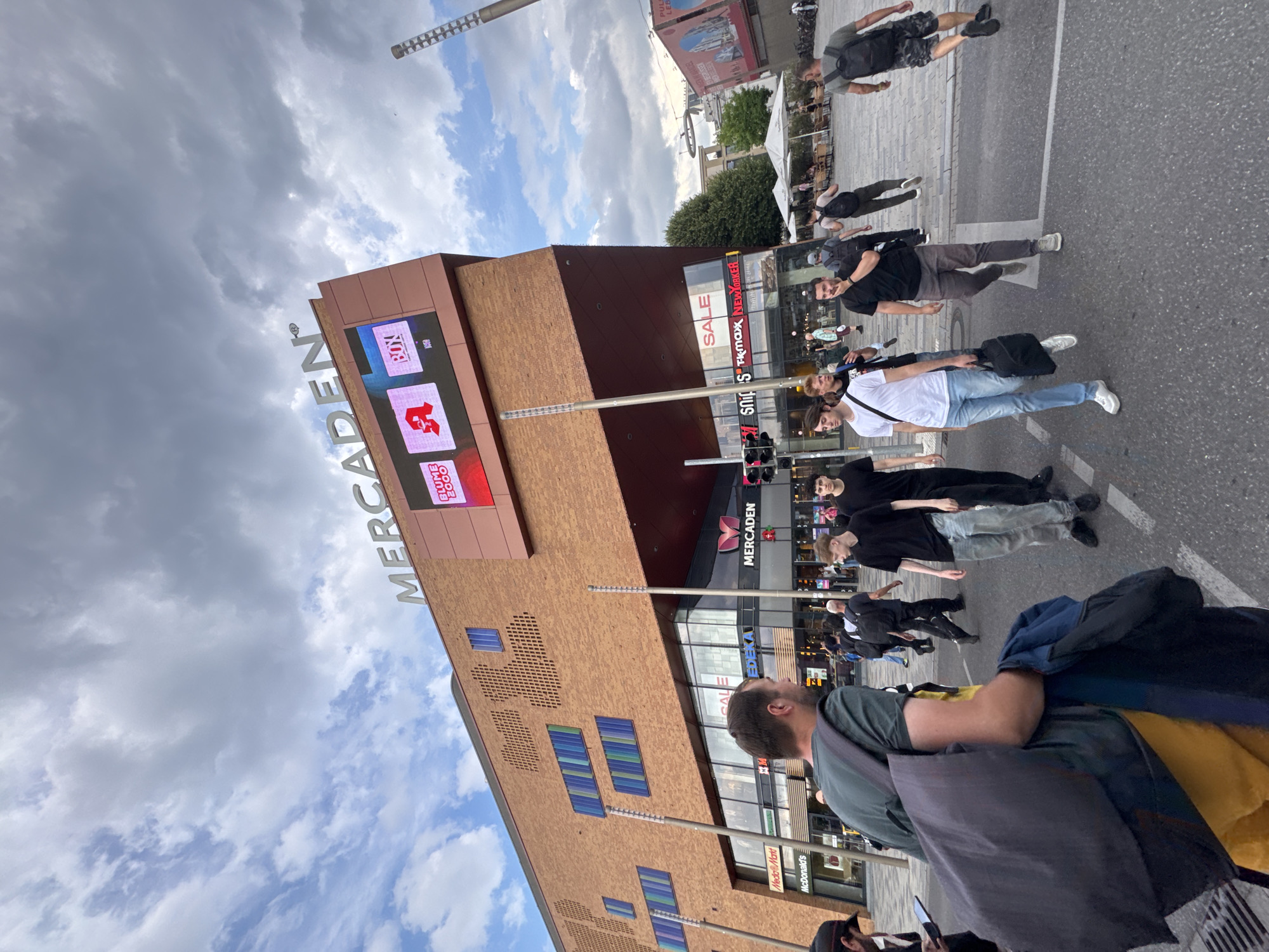 This is a portrait-oriented, eye-level, outdoor photograph taken on a cloudy day. The scene appears to be a bustling urban environment, likely a shopping district or pedestrian zone, in Böblingen, Germany. Dominating the frame is the exterior of a large, modern building with a brick facade, identified by the prominent signage "MERCADEN". Below the "MERCADEN" sign, a large digital display shows various advertisements, including "BOX" and a red logo with white text. Other visible commercial establishments include "Media Markt", "McDonald's", and "Temax NEWYORKER", suggesting a retail hub.
The foreground features several individuals, mostly men, walking or standing. Their clothing suggests casual attire, and some are carrying bags or backpacks. The ground is paved, indicating a pedestrian area or street. The people are oriented in various directions, with some looking at the camera and others looking away. The overall mood appears to be lively and active, typical of a public space with ongoing commerce and pedestrian traffic. The time of day is difficult to ascertain precisely due to the cloud cover, but the lighting suggests it is daytime.