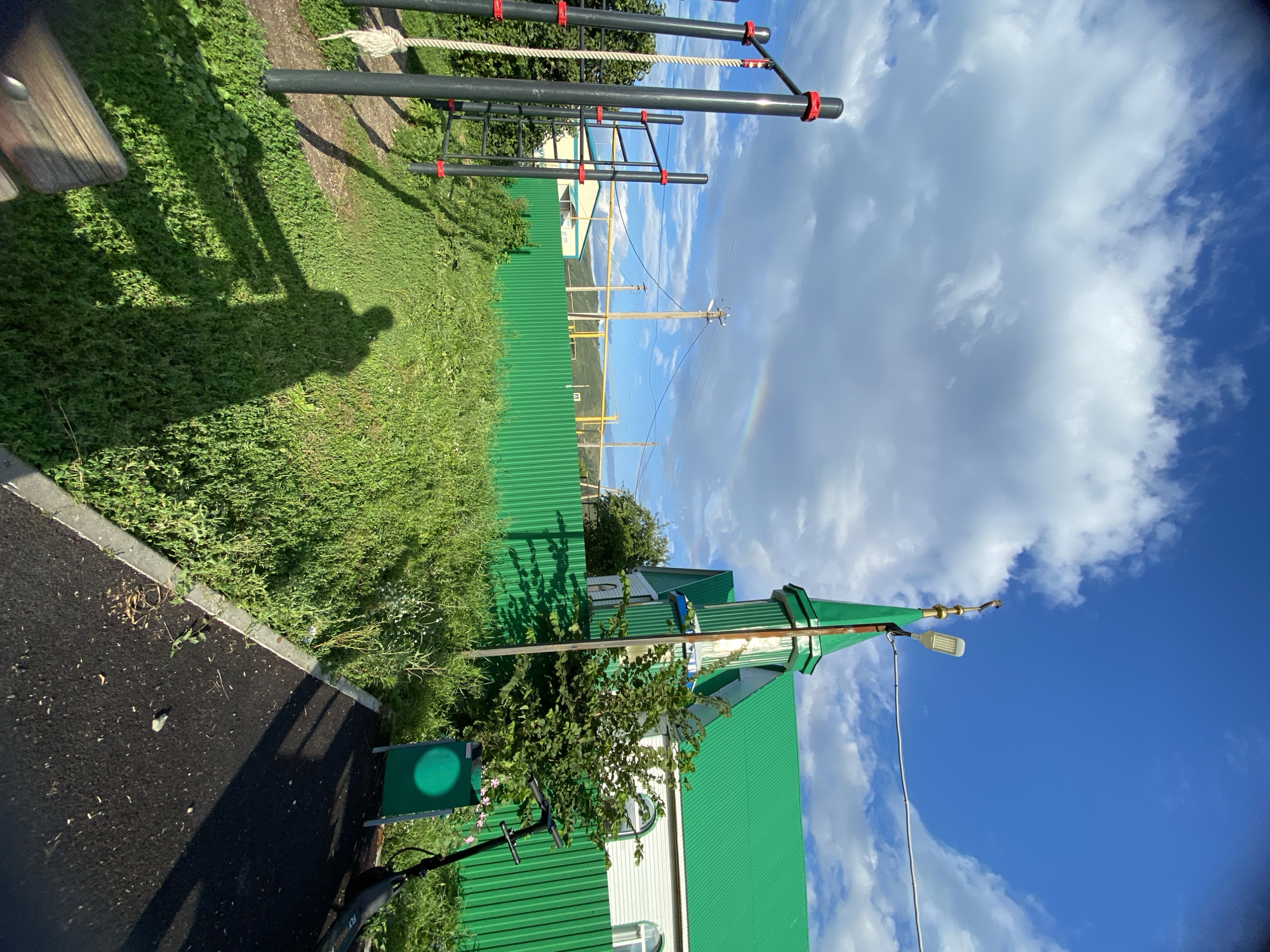The image depicts an outdoor scene with a green corrugated metal fence in the foreground, separating a grassy area from a paved path. To the left, a black metal outdoor exercise rig with red accents is visible, featuring a rope and horizontal bars. A long shadow of a person, presumably the photographer, is cast across the grass.
In the background, a building with a vibrant green roof and a pointed spire is prominent. The building has white siding and arched windows. A streetlamp is attached to a pole near the building. A faint rainbow is visible in the sky above and to the left of the building.
The environment appears to be a public space, possibly a park or a schoolyard, given the exercise equipment and the presence of a scooter parked near the fence. The sky is a bright blue with scattered white clouds, suggesting fair weather. The overall setting is likely residential, as indicated by the houses and power lines in the distance.
The location is identified as Maloarslangulovo, Russia.