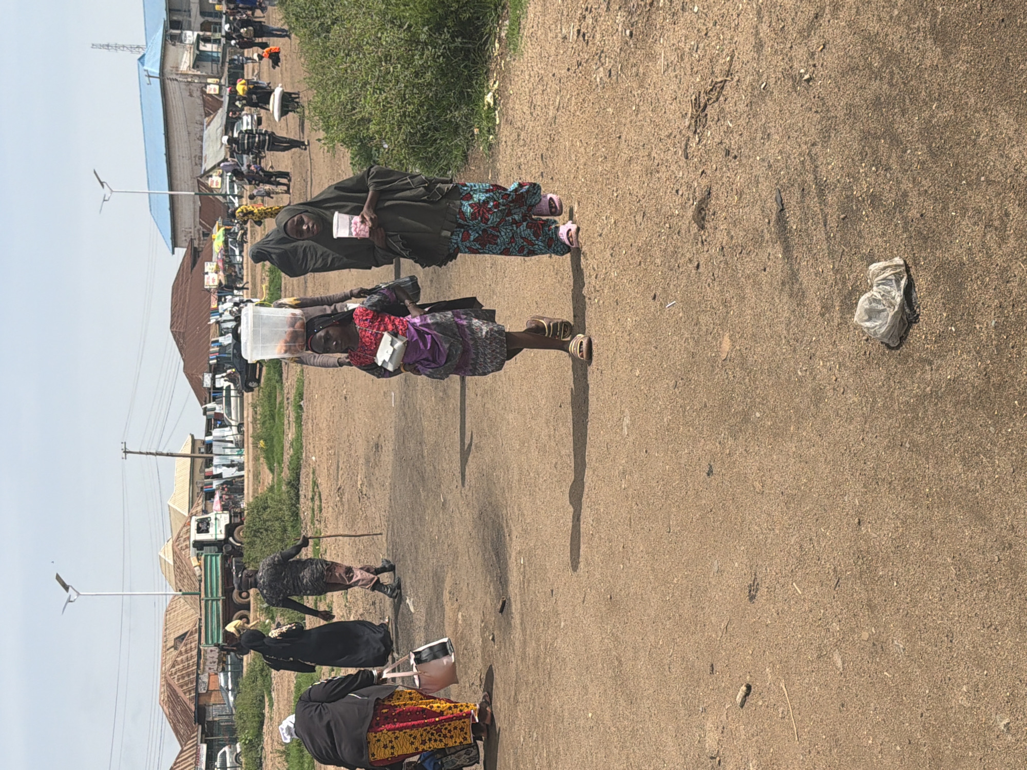 The image depicts a street scene in Bukuru, Nigeria. In the foreground, two young girls are walking. The girl on the left, wearing a colorful dress and a white purse, carries a large clear plastic container filled with fruit on her head. The girl on the right, wearing a dark hijab and a patterned skirt, holds a cup filled with pink items. Behind them, other people are walking and interacting in a bustling outdoor market environment. Various stalls and buildings line the background, suggesting a commercial area. A truck is parked in the distance, and overhead power lines are visible. The ground is dusty and uneven, typical of a developing urban setting. The overall impression is one of everyday activity in a Nigerian market.