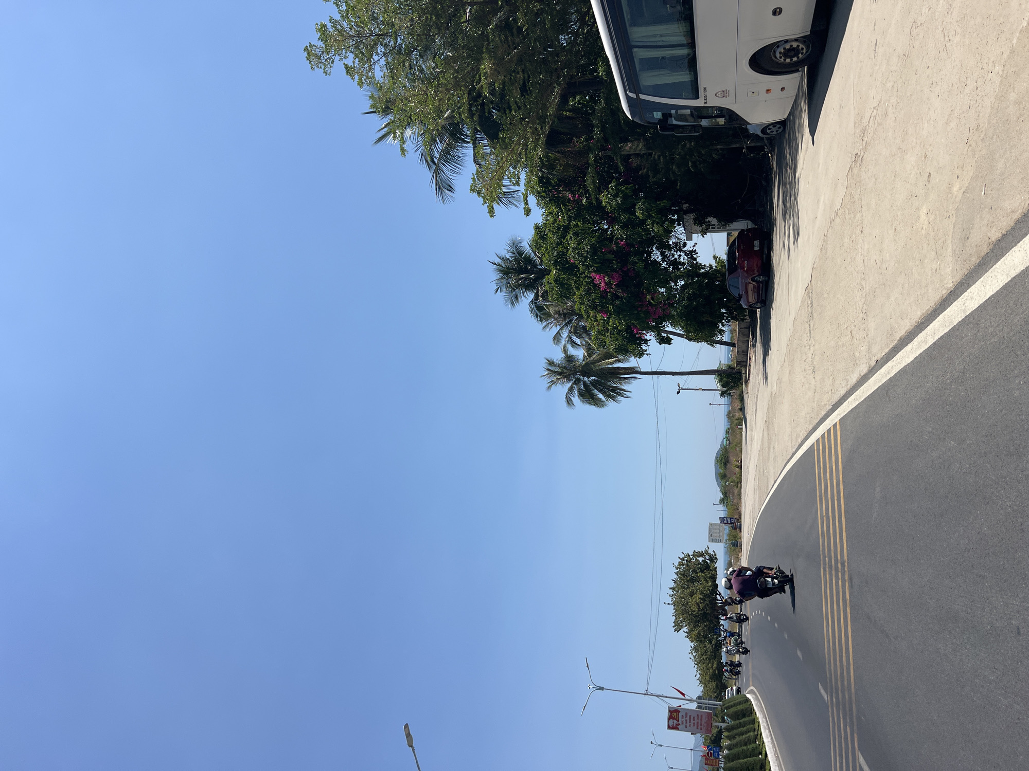 The image shows a street scene in Nha Trang, Vietnam. A road curves from lower right to upper right, with a solid white line on the right edge and dashed yellow lines dividing the lanes. Several motorcycles are riding on the road, moving away from the viewer. The sky is clear and blue. Along the upper right edge of the road is a concrete sidewalk, where a white bus is parked, followed by a red car. Trees, including palm trees, are located on the left edge of the road. Streetlights are visible along the road. A glimpse of the ocean is visible in the distance, near the center. The road is asphalt, showing signs of wear. The scene is bathed in sunlight.