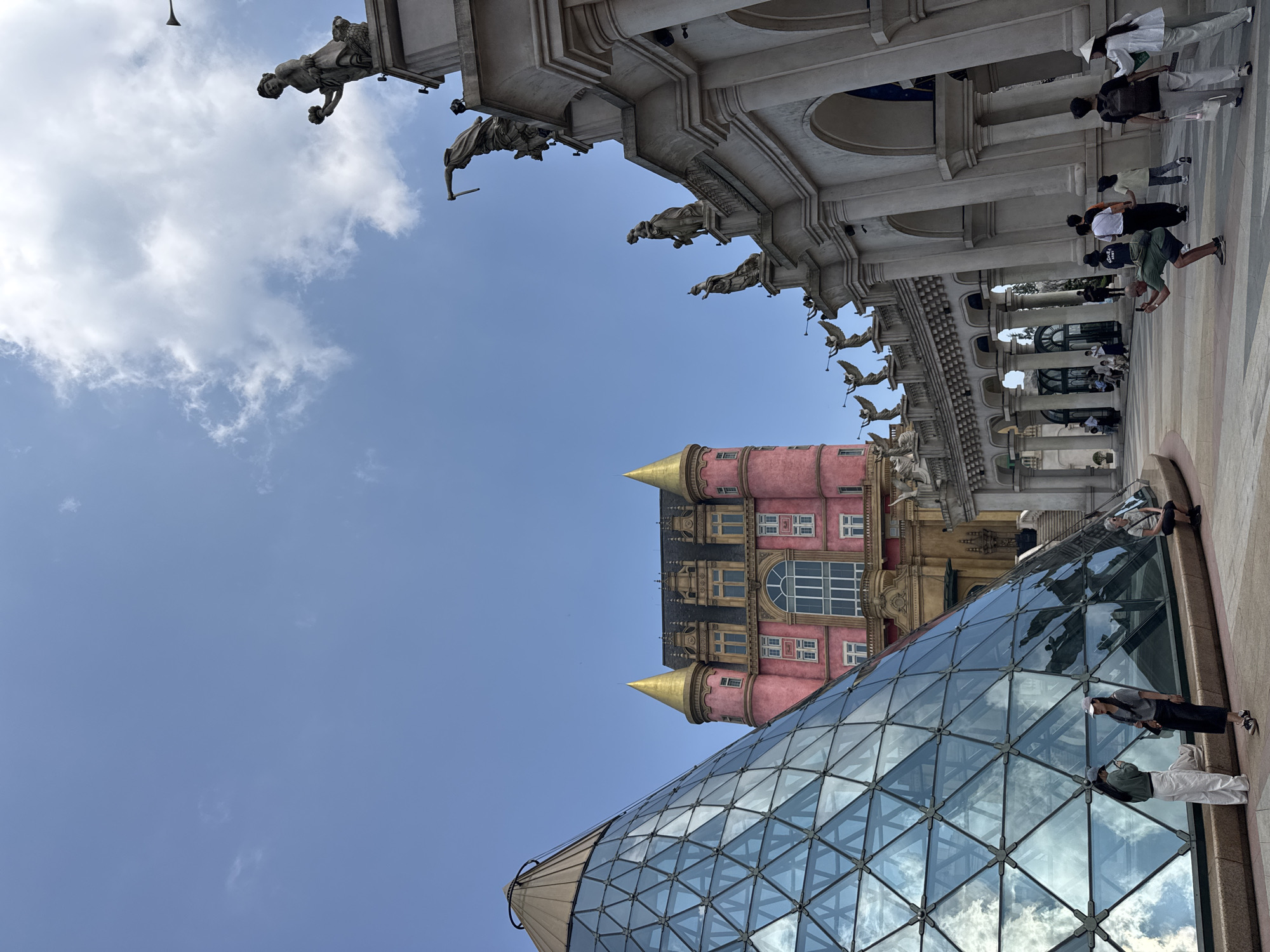 The image captures an outdoor plaza at Sun World Ba Na Hills in Đà Nẵng, Vietnam, under a clear blue sky with sparse white clouds. To the left, a large, angular pyramid structure constructed from numerous triangular glass panels and capped with a bronze apex reflects the sky and ambient light. Two individuals stand near the base of this pyramid; one in a green long-sleeved shirt and white pants, and another in a grey t-shirt and dark skirt, both appearing to engage with mobile devices. A third individual sits on a low retaining wall to the right of the pyramid base.
In the midground, centrally positioned, a prominent multi-story building with a distinct pink facade, a dark roof, and golden conical turrets rises. It features multiple windows and intricate architectural detailing. To the right of the frame, a long, neoclassical-style structure with a series of arched openings supported by columns forms a colonnade. Its upper ledges and roofline are adorned with numerous grey stone statues depicting human figures, some holding trumpets, others in various classical poses.
The plaza ground is paved with light-colored, geometrically patterned tiles. Multiple individuals traverse the plaza. One person in a green t-shirt and dark shorts is observed in a crouched position, holding a mobile phone horizontally, appearing to take a photograph. Other pedestrians are visible walking in various directions across the plaza; one individual wears a traditional conical hat (nón lá), and another carries a backpack. The scene is illuminated by bright daylight.