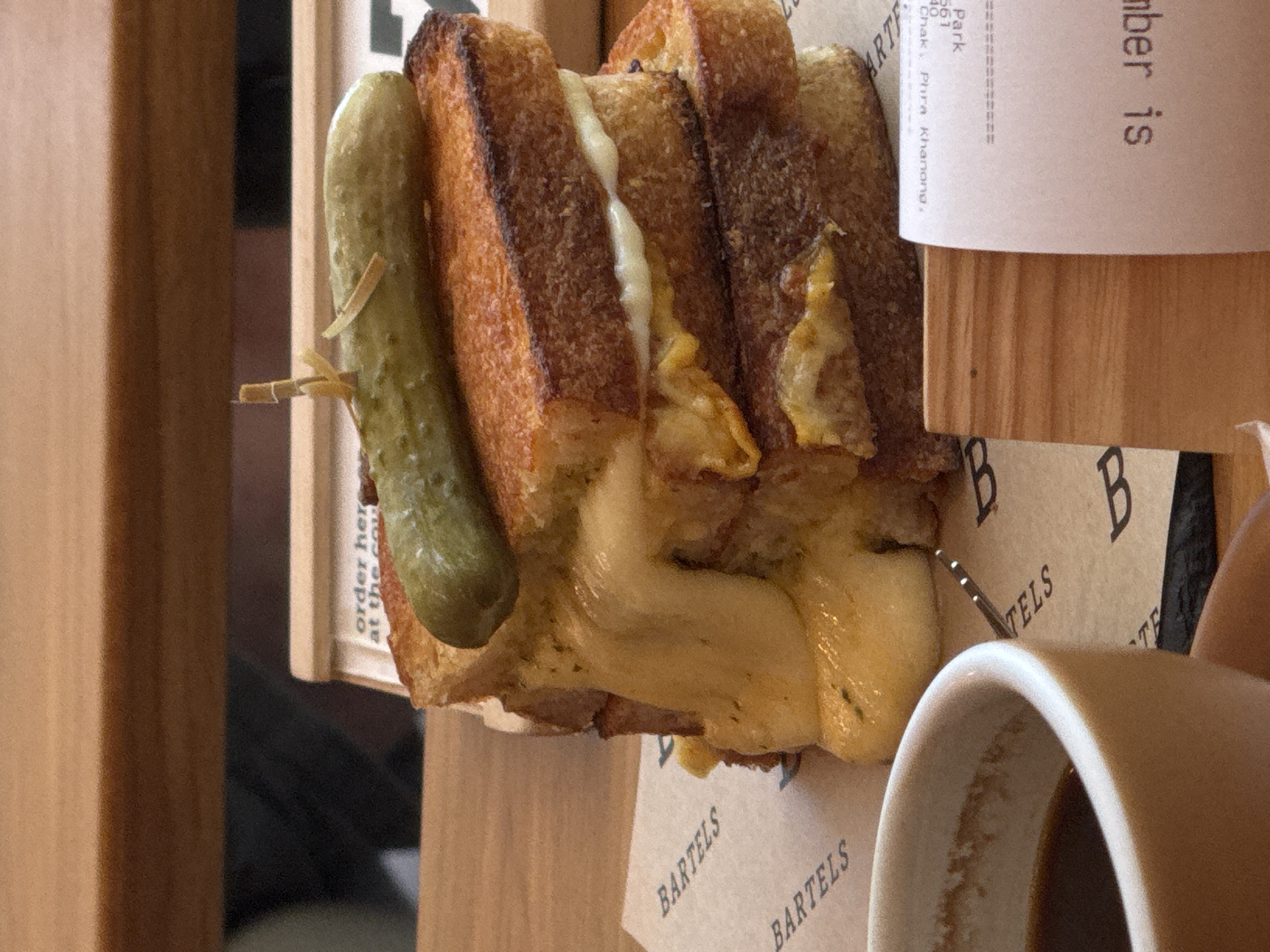 The image shows a close-up of a grilled cheese sandwich, topped with a pickle and held together with a toothpick. The sandwich is served on a brown paper liner with the logo "BARTELS" printed repeatedly. A cup of dark liquid, likely coffee, is partially visible in the lower left corner. In the background, there is a sign that reads "order here at the counter". A receipt with text in a foreign language, including "Park" and "Chak, Phra Khanong", is visible to the right, suggesting the location is in the Phra Khanong area of Bangkok, Thailand. The overall environment appears to be a casual dining setting with wooden furniture. No people are directly visible in the frame.