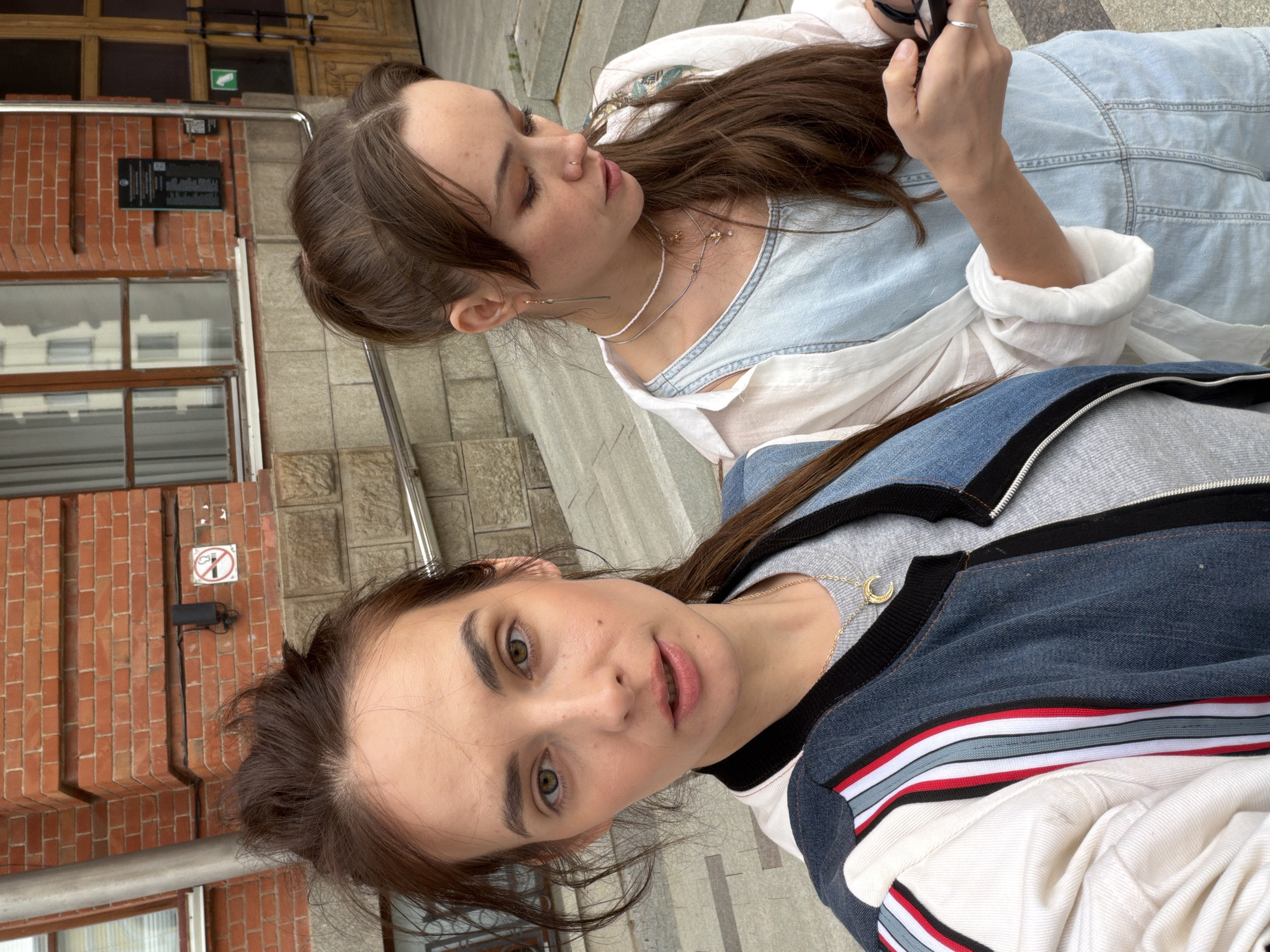 Two young women are posing for a selfie in front of a red brick building, likely in the city of Ufa, Russia. The woman in the foreground is closer to the camera and has her hair pulled up in a messy bun. She is wearing a denim jacket with white, red, and blue stripes on the sleeves and a grey t-shirt. The woman in the background is looking down at her phone and has her hair down in loose waves. She is wearing a light blue denim jumpsuit and a white open shirt over it. She has several necklaces on. The building behind them has large windows and a wooden door. There are also stairs leading up to the building. A no-smoking sign is visible on the brick wall to the left.