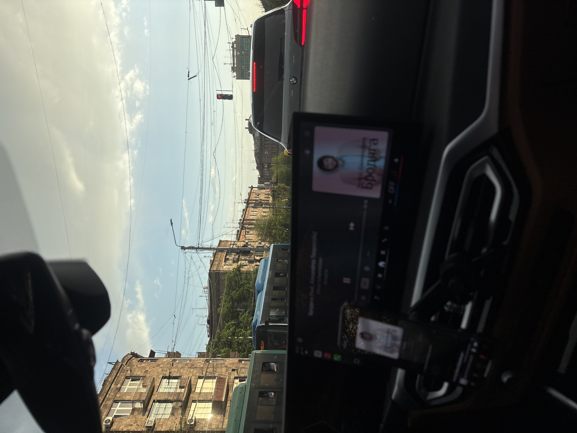 The image depicts a view from inside a car, likely a BMW, stopped in traffic in Yerevan, Armenia. Through the windshield, buildings line the street. Above, a cloudy sky is crossed by power lines. A red traffic light is visible. Buses are near the car in traffic. Inside the car, the dashboard screen displays a person and text in Armenian reading "Бролига". A smartphone mounted on the dashboard also shows a display.