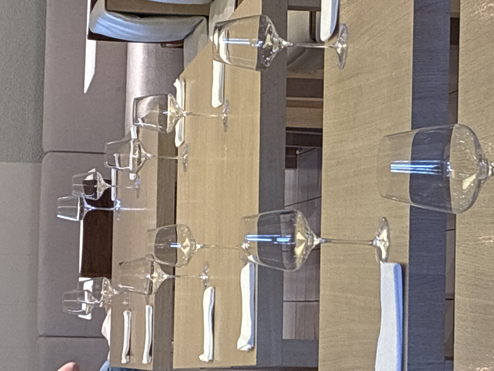 The image depicts a row of set tables, likely in a restaurant, with wine glasses and napkins arranged for diners. The location is possibly in Belgrade, Serbia, although this cannot be verified from the visual information provided. The glasses are transparent and clean, implying readiness for service. The napkins appear white and neatly folded. The tables have a light brown, wooden surface. The background shows additional tables and chairs, along with sections of upholstered furniture, suggesting a sophisticated dining setting. The image is taken at an angle, providing a perspective view of the table arrangement.