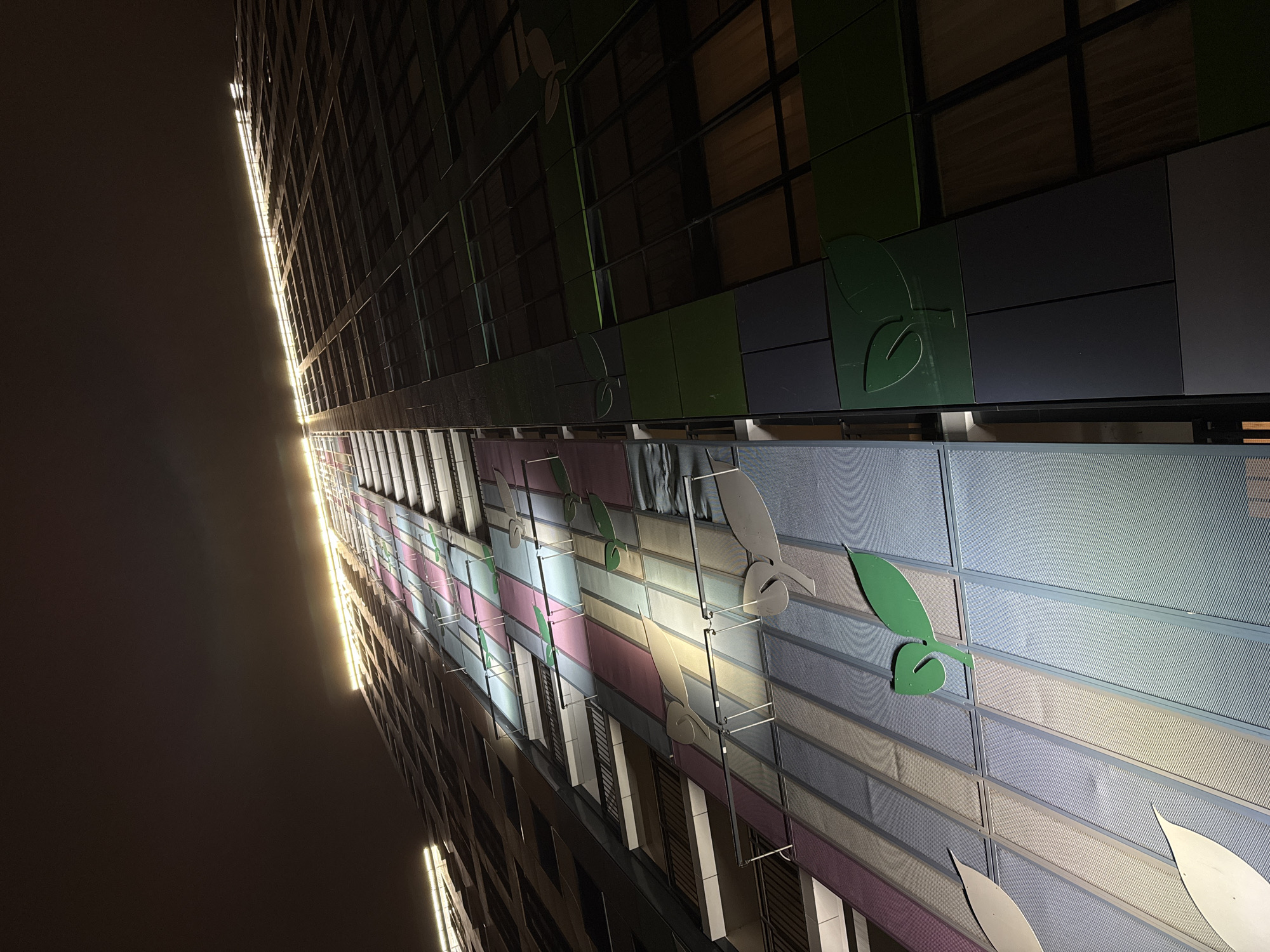 An upward-angled view captures the illuminated facade of a tall, modern building at night in Saint Petersburg, Russia. The image emphasizes the building's intricate and colorful architectural details.
The building's lower and mid-sections feature a vibrant facade composed of rectangular panels in various pastel shades, including light blue, pink, and yellow, arranged in horizontal stripes. These panels appear to have a textured, mesh-like surface. Interspersed among these colorful sections are decorative leaf-shaped elements, primarily in green, but also in light grey or white, adding an organic motif to the geometric design.
Above this colorful section, the building transitions to a darker facade with panels in shades of dark green and grey, also adorned with similar green leaf designs. Windows are visible in this upper section, reflecting some light.
A striking, intensely bright, vertical line of light runs along the far left edge of the building, creating a stark contrast against the dark night sky. This strong illumination suggests powerful external lighting or internal lights creating a distinct beam. The light creates reflections on the adjacent surfaces of the building. The overall scene is devoid of people or overt activity, focusing solely on the building's architectural presence and nocturnal lighting. No visible text is present in the image.