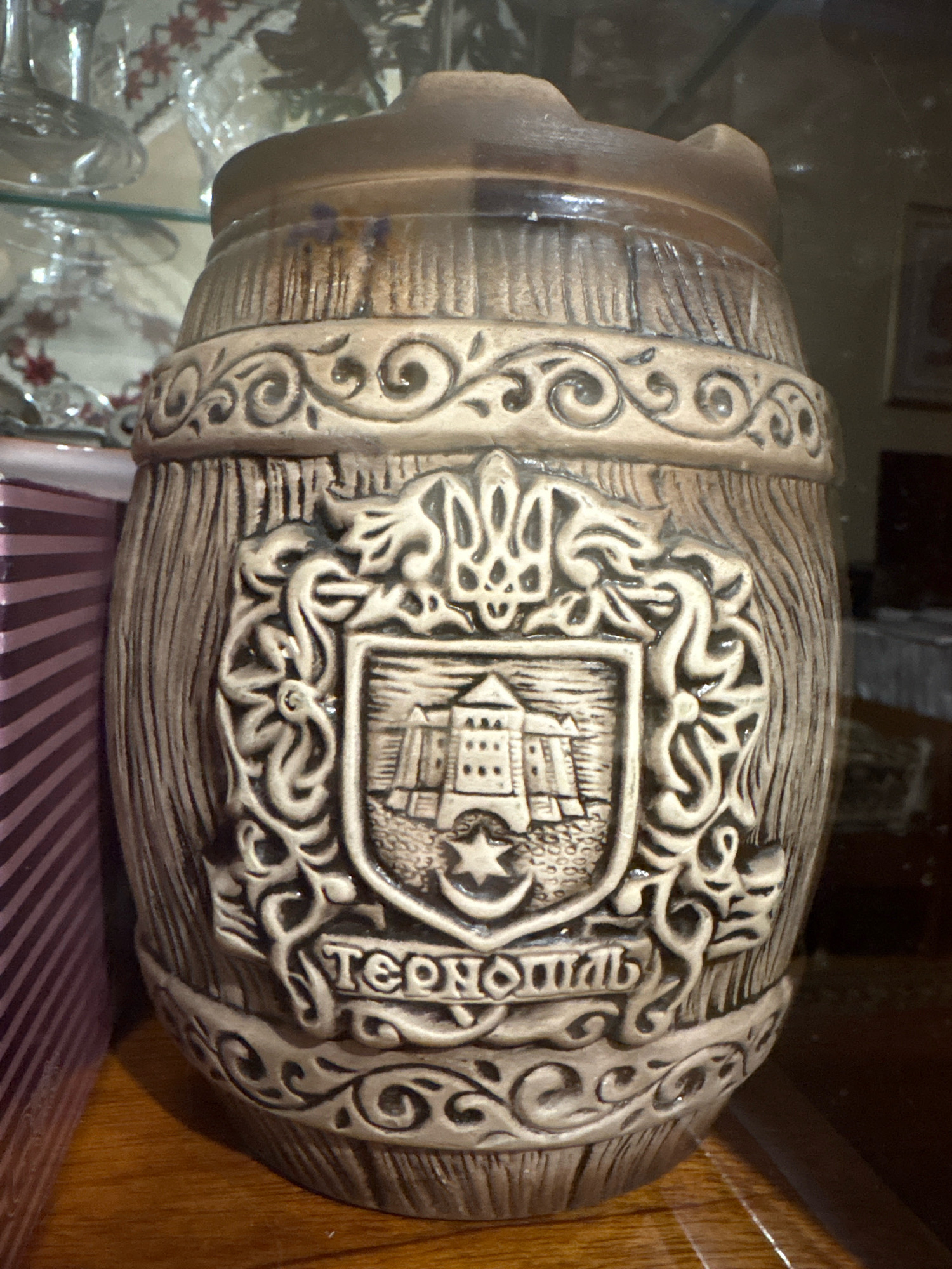 The image shows a close-up of a ceramic barrel with an embossed shield and Ukrainian inscriptions. The barrel has a distressed wood-like texture and decorative scrollwork around its circumference. The central shield depicts a fortress or castle with a star below it. Above the shield is a stylized emblem resembling a trident. The inscription below the shield reads "ТЕРНОПІЛЬ" (Ternopil). The barrel is placed on a wooden surface, likely a table or shelf, and is partially obscured by a striped object on the left. The background is out of focus and appears to be an indoor setting with glass shelves and other objects, suggesting it might be a display cabinet. The lighting is dim, casting shadows on the barrel. The image is taken from a slightly low angle, focusing on the details of the ceramic craftsmanship.
