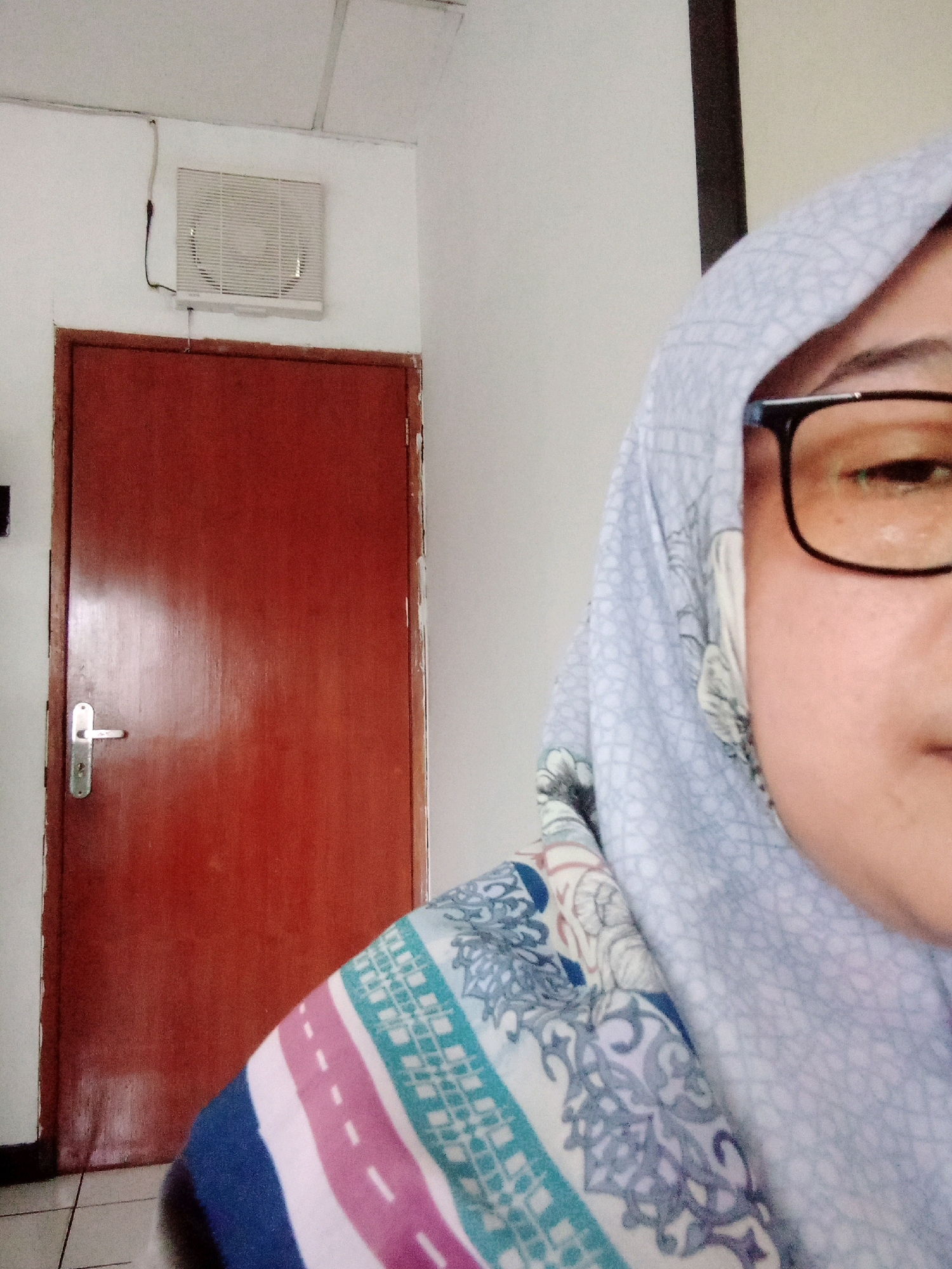 In a room, possibly a residential or office space, a woman wearing a hijab and glasses is captured in a close-up, half-profile selfie. The background reveals a closed, dark reddish-brown wooden door with a silver handle and lock mechanism, to the left of the woman. Above the door, on a white wall, a white rectangular ventilation grille is mounted. The overall environment suggests an indoor setting, likely within Balaraja, Indonesia, given the provided context. The woman's attire includes a light blue hijab with a subtle pattern and a garment with stripes of pink, white, and teal, adorned with intricate blue and white floral designs.