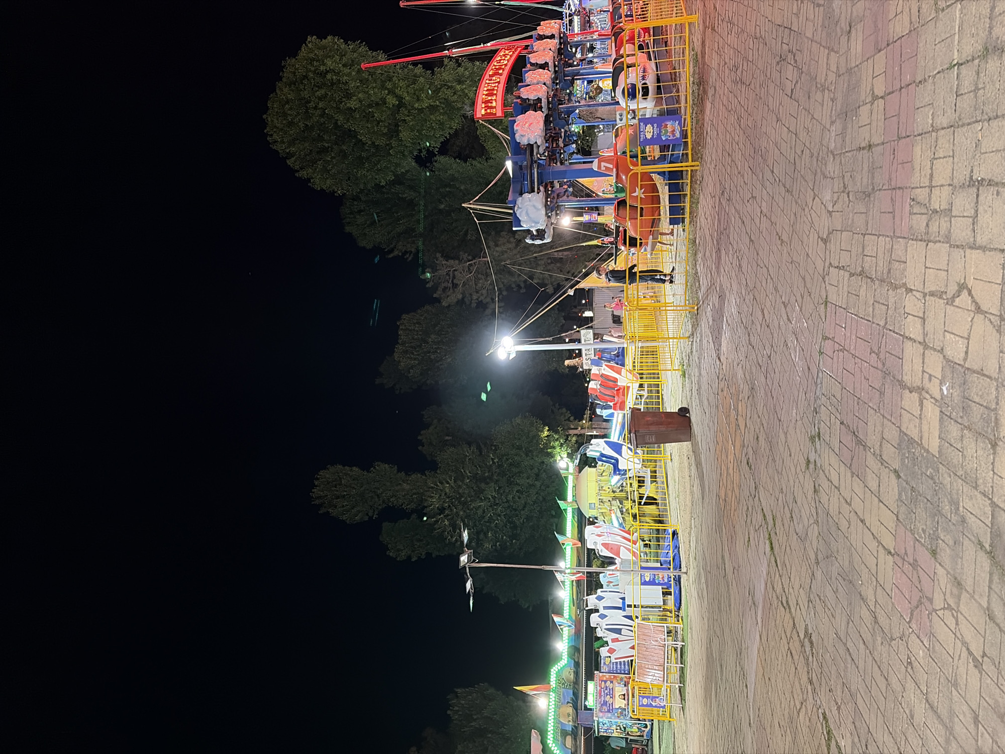 An outdoor amusement park is captured at night. The foreground features a brick-paved walkway, with amusement rides visible in the mid-ground. These rides include a "Flying Tiger" ride with red and blue carriages shaped like planes and clouds, and a smaller ride with white and red seats arranged in a circular pattern. Yellow safety barriers surround some of the rides. In the background, several tall trees create a dark silhouette against the night sky, with some areas illuminated by amusement park lighting. A few people are present, with one individual standing near the "Flying Tiger" ride. The overall scene depicts a lively, albeit quiet, amusement park at night.