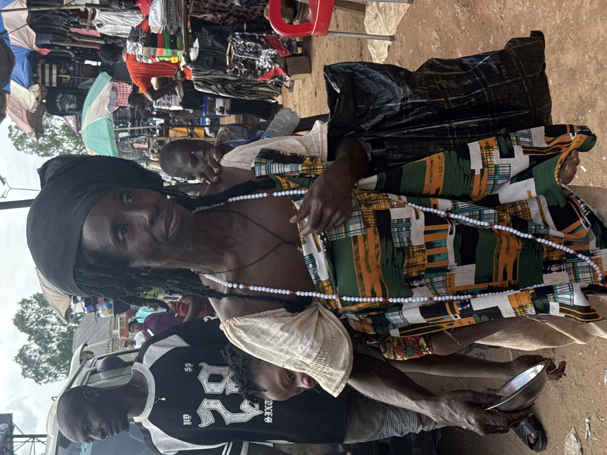 A woman wearing a black headwrap and a colorful patterned wrap is holding a baby in a sling. She is standing in an outdoor market in Onitsha, Nigeria. To her left, a man in a black and white shirt with the number "28" on it is looking to his left. In the background, there are market stalls with clothing and other goods, as well as several people. A red plastic chair is visible to the right. The ground is dirt.
