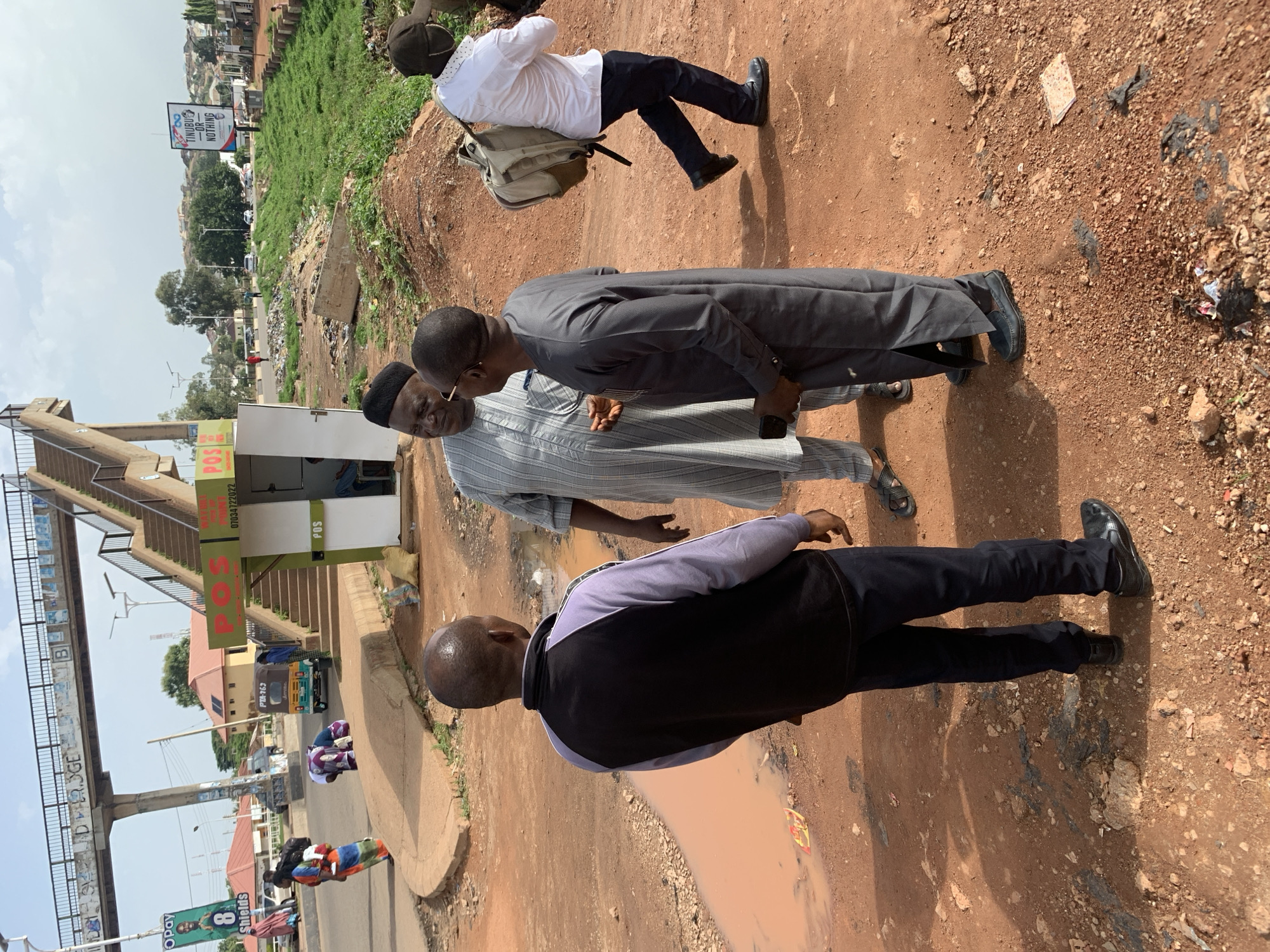 In the city of Gura Top, Nigeria, an outdoor scene unfolds near a pedestrian bridge. Three men are engaged in conversation on a dirt road. The man on the left, wearing a black and purple jacket over black pants, stands with his back to the camera. The man in the center, dressed in a light-colored striped robe and a black cap, faces the man on the right. The man on the right, in a dark grey robe and black cap, holds a phone in his left hand. Behind them, a white structure with "POS" signage and stairs leading up to a pedestrian bridge suggests a public thoroughfare. Further in the background, a tuk-tuk, several vehicles, and buildings are visible, indicating a semi-urban environment. A person in a white shirt and dark pants with a backpack walks away from the group towards the right of the frame. The ground is a mix of reddish-brown dirt, gravel, and a muddy puddle, with scattered debris. Lush green vegetation covers a slope in the background, and a billboard with the slogan "TINUBU OR NOTHING" is visible on the horizon.