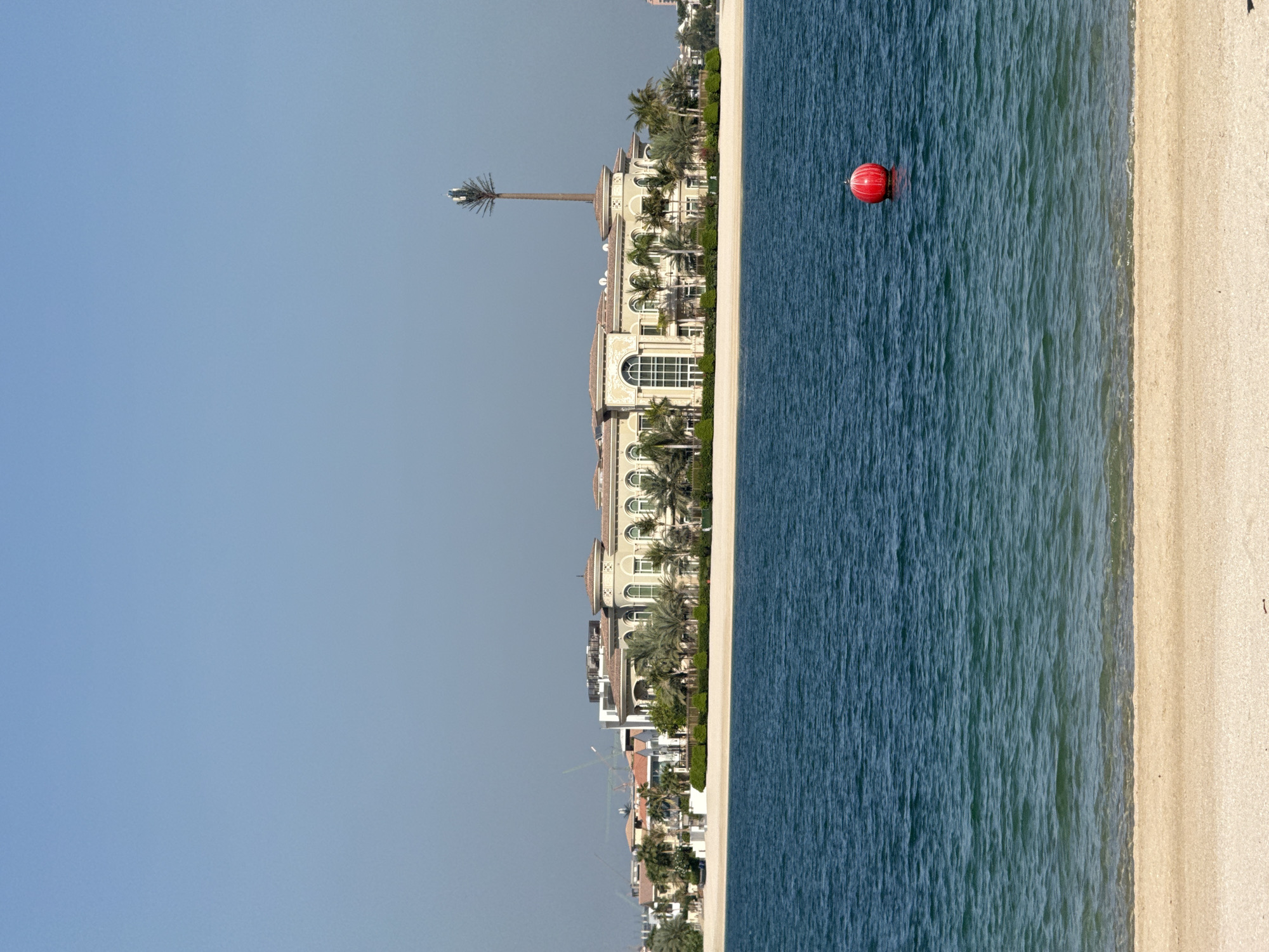 The image shows a view of a large, opulent villa situated on a sandy beach with palm trees in front of it. The villa has a grand facade with arched windows and a domed roof. A tall, slender tower resembling a palm tree with antennas at the top stands behind the villa. In the foreground, a calm blue sea laps gently against the sandy shore. A single bright red buoy floats in the water. The sky is clear and blue. Construction cranes are visible in the distance, suggesting ongoing development in the area. This scene is characteristic of the man-made islands of Dubai, specifically the Palm Jumeirah, known for its luxurious villas and waterfront properties.