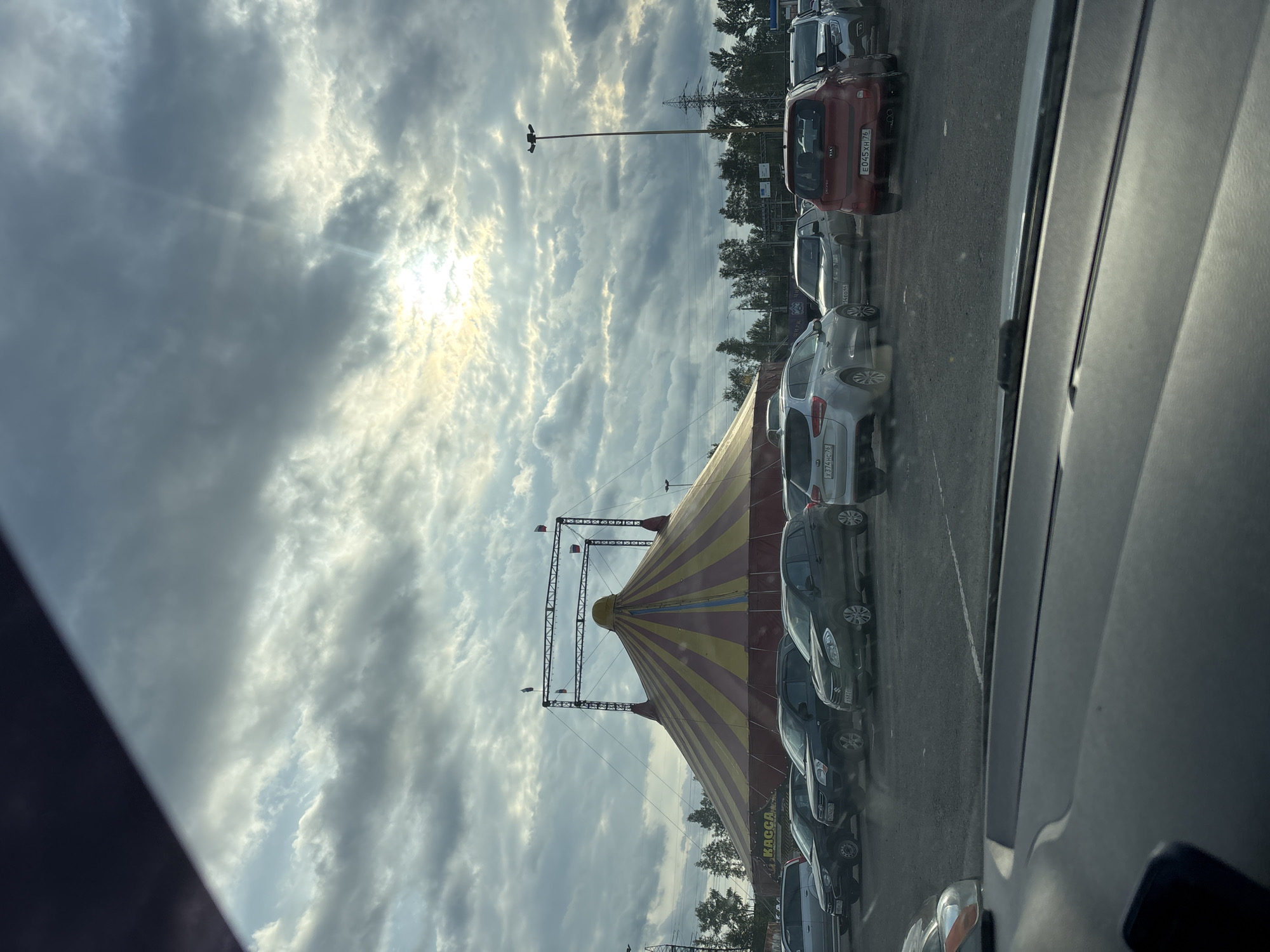 The image depicts an outdoor parking lot with a large, striped circus tent in the background. The tent is predominantly yellow and red, with a peaked roof supported by a metal structure. The word "КАССА" (KASSA), meaning "ticket office," is visible on the side of the tent. Several cars are parked in the lot, including a red Kia, a silver wagon, and various other sedans and SUVs. The sky is cloudy with rays of sunlight breaking through, suggesting it is either sunrise or sunset. The environment appears to be a flat, paved area with some trees and power lines visible in the distance, characteristic of a semi-urban or suburban setting. The perspective suggests the photo was taken from inside a vehicle. Based on the license plates visible on some of the cars, which appear to have Russian alphanumeric characters and regional codes like "76", the location is likely Korobovo, Russia.