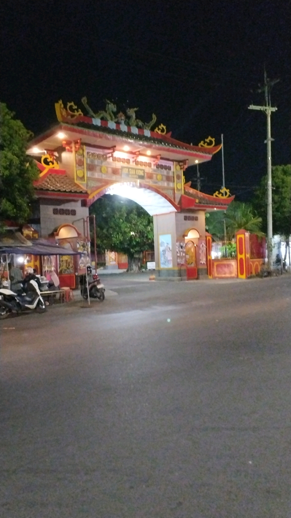 A nighttime street scene shows a brightly lit, ornate gateway with traditional East Asian architecture. The gateway, which appears to be an entrance to a temple or cultural site, is adorned with intricate carvings and decorative elements, including dragon figures along the roofline. The gateway is illuminated by various lights, highlighting its colorful details.
To the left of the gateway, there is a small, covered structure that appears to be a food stall or a small shop. A white scooter and a dark-colored scooter are parked in front of this stall, with two individuals seated at a table.
The gateway itself is wide and arched, allowing passage through to an area with trees visible in the background. The area behind the gateway features more traditional architectural elements and brightly colored walls.
To the right of the gateway, a utility pole with wires is visible, along with trees. The ground in the foreground is paved asphalt, indicating a road or street. The overall atmosphere is that of an active evening in a place with a strong cultural identity. The location is identified as Purwotengah, Indonesia.