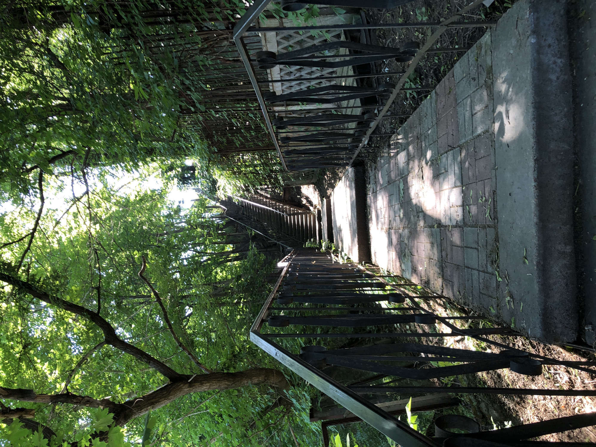 An outdoor, low-angle shot captures a brick pathway leading up a steep staircase lined with lush green trees and foliage. The pathway is partially shaded by the sunlight filtering through the leaves, creating dappled patterns on the brickwork. A dark metal railing runs along the right side of the pathway, and another railing is visible on the left, leading up the stairs. The stairs themselves appear to be made of concrete or stone, with brick risers. The environment suggests a park or a wooded area within a city, possibly a scenic overlook or a path through a steep incline. There are no people visible in the frame.