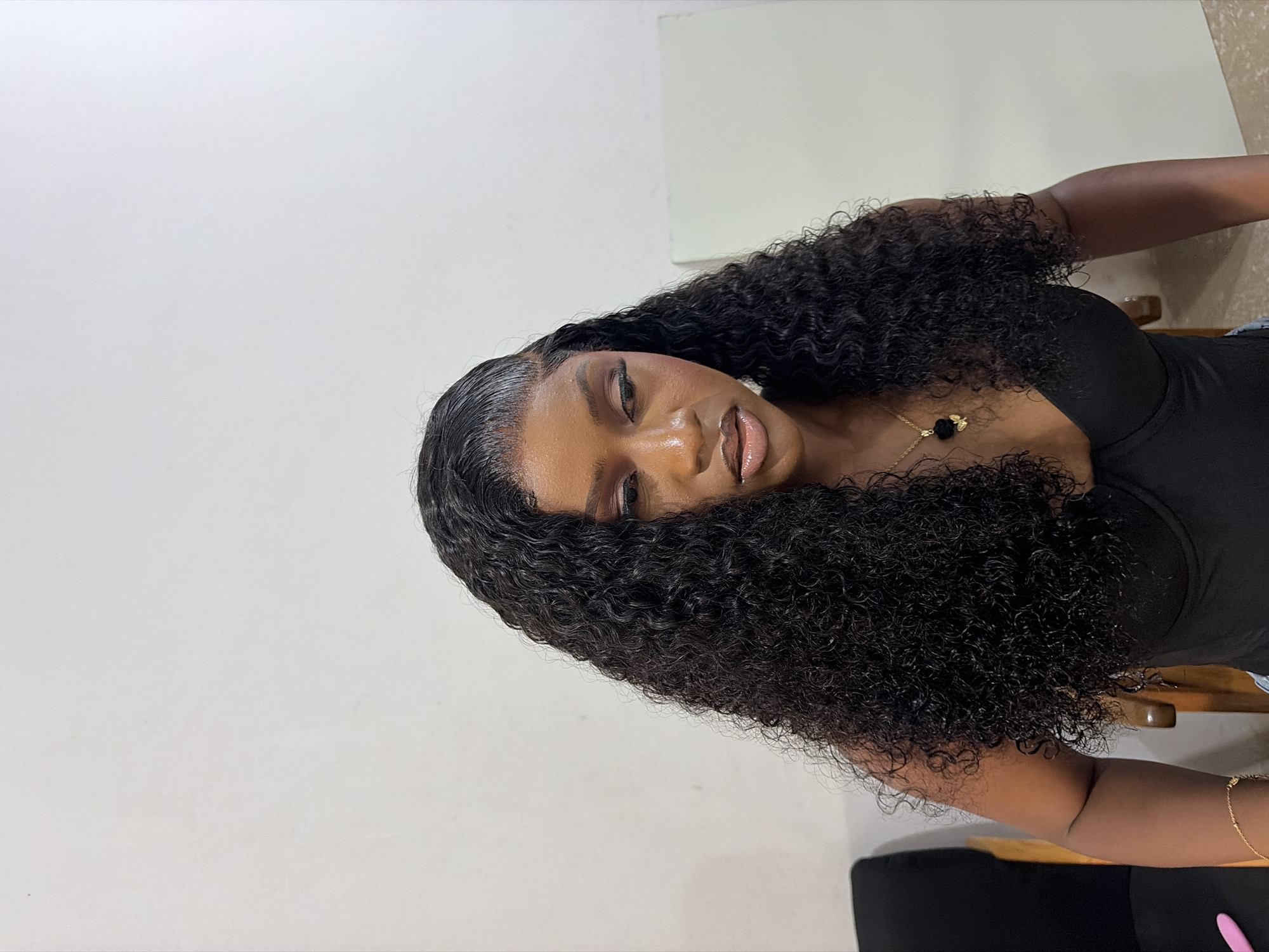 A Black female is depicted seated indoors, positioned from the chest up against a plain, light-colored wall. She has long, dark, curly hair with a side part, styled to lay flat at the hairline. Her facial makeup includes defined eyebrows, eyeshadow, eyeliner, and nude-toned lipstick. Her gaze is directed downwards and to the left, conveying a neutral expression. She wears a black bustier-style top, a gold chain necklace with a black, possibly floral or butterfly-shaped pendant, and a gold chain bracelet on her right wrist. A dark chair with visible wooden elements is partially present on the left side of the frame, and a white rectangular panel stands on the right in the background. The environment appears to be artificially lit.