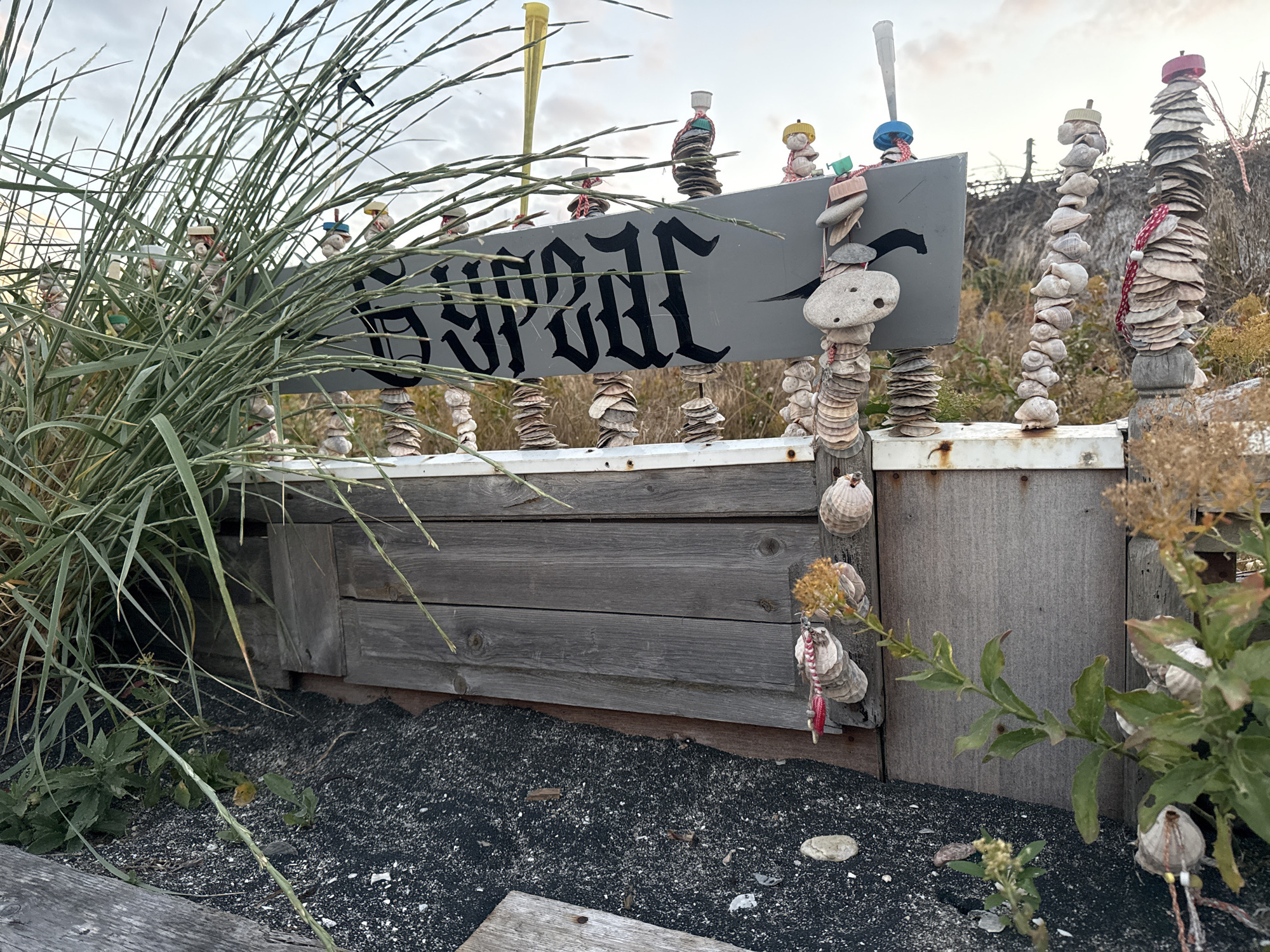 The image shows a wooden fence with a gray sign that has black gothic lettering. The sign appears to say "BURGASS". The fence is decorated with stacks of shells and stones, some of which have small figures or hats on top. In front of the fence is dark sand and some sparse vegetation. To the left, there is a large clump of tall grass. The background shows a cloudy sky and dry brush. The scene is outdoors, likely near the sea, given the abundance of shells. The sign suggests the location is Burgas, Bulgaria.