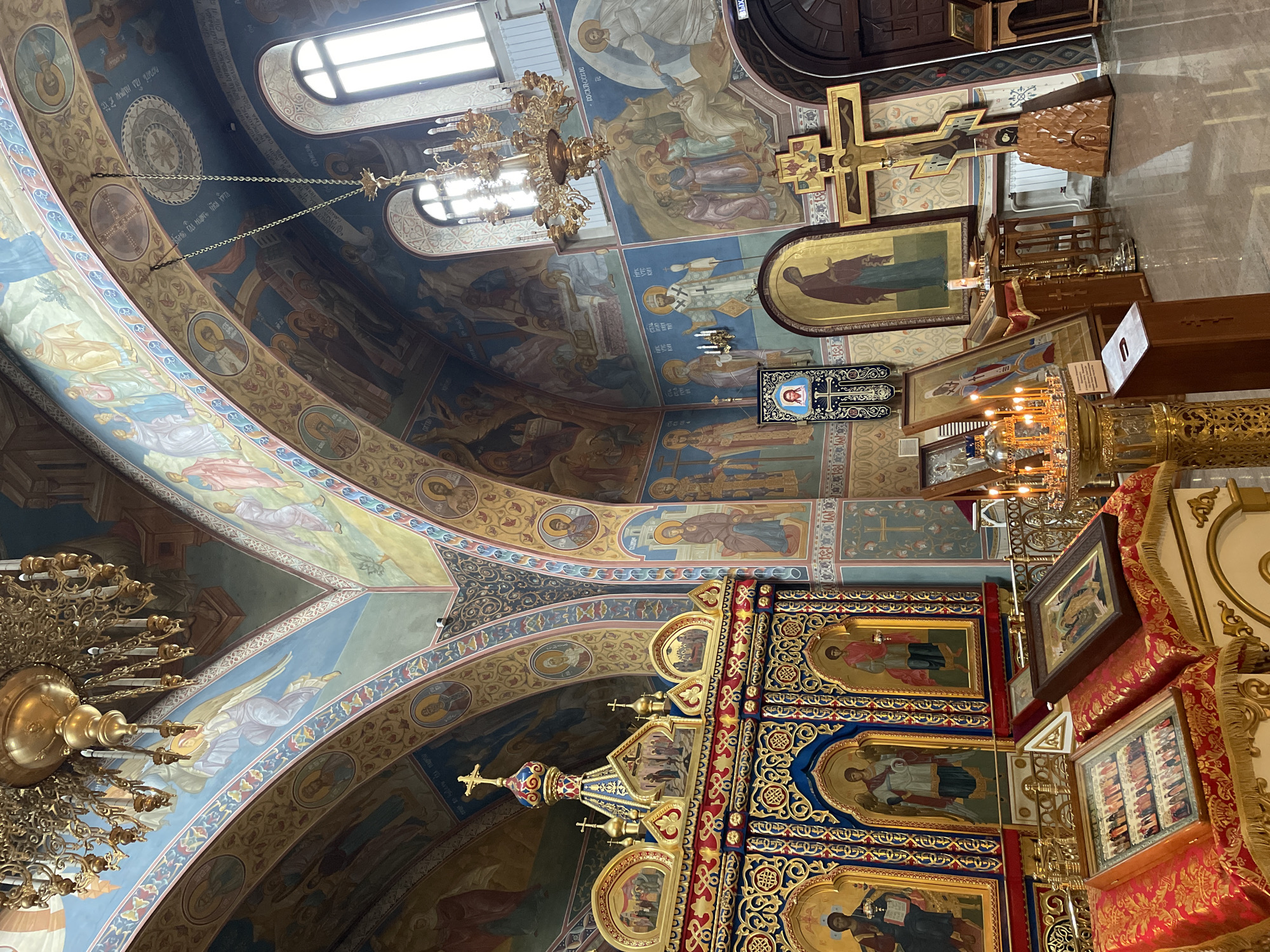 The image captures the interior of a richly decorated Orthodox Christian church in Krasnoyarsk, Russia. The focus is on the ceiling and the upper portions of the walls, adorned with religious frescoes depicting various biblical scenes and saints. The paintings are executed in a traditional style with bright colors and ornate borders.
A large, gilded chandelier hangs from the ceiling, adding to the opulence of the space. Religious icons, framed in gold, are visible on the walls. A large wooden cross with a depiction of Christ is prominently displayed. The lower portion of the image reveals elements of the iconostasis, a screen separating the nave from the sanctuary, decorated with intricate carvings and religious images. The overall impression is of a sacred space with a strong sense of historical and artistic significance.