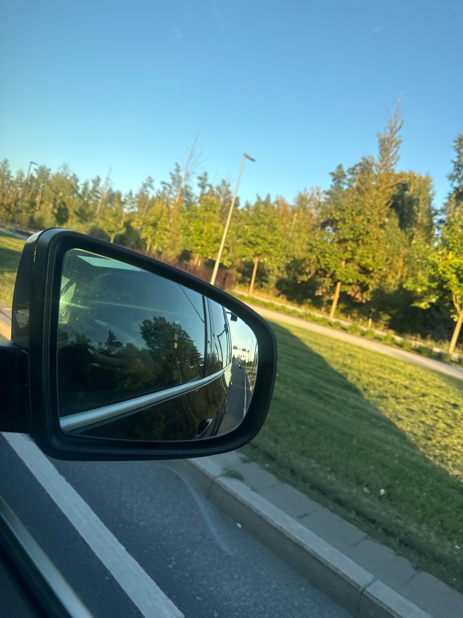The image captures a view from a moving vehicle, looking towards the right side mirror and the environment outside. The right side mirror reflects the interior of the car, showing a person's hands on the steering wheel, suggesting the vehicle is in motion. The exterior view shows a roadside with a well-maintained grassy slope and a line of trees. A street lamp is visible in the distance. The sky is clear and blue, indicating daytime. The presence of a street lamp and the overall urban-like landscaping suggests this is likely a suburban or well-developed area within a city, possibly on the outskirts of Tallinn, Estonia, given the prompt's context. The road markings in the foreground indicate a paved surface. The lighting suggests it is either early morning or late afternoon, with the sun casting long shadows.