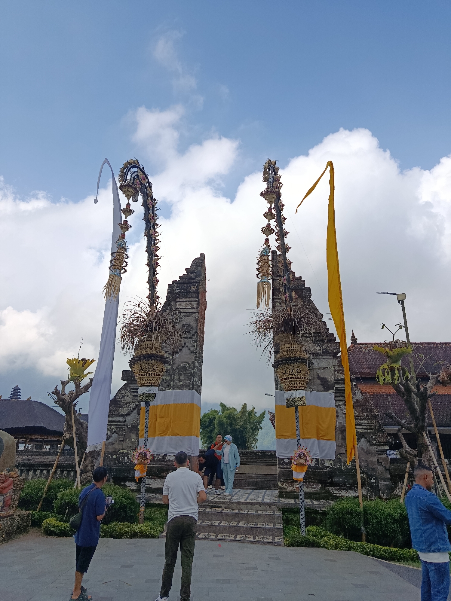 In the city of Baturiti, Indonesia, a stone gate with elaborate carvings stands open under a cloudy sky. The gate is flanked by tall, ornate poles, one adorned with a white fabric banner and the other with a yellow flag. People are seen in the foreground and on the steps leading through the gate. One person in the foreground, wearing a blue shirt and dark shorts, is looking down at a phone. Another person, in a white shirt and green pants, stands with their back to the camera. A group of people in lighter clothing are visible on the steps, one person in a light blue outfit. Lush green bushes and trees surround the area, and traditional Balinese architecture, including a tiled roof building, is visible in the background. The overall scene depicts a cultural site, likely a temple, with visitors present.