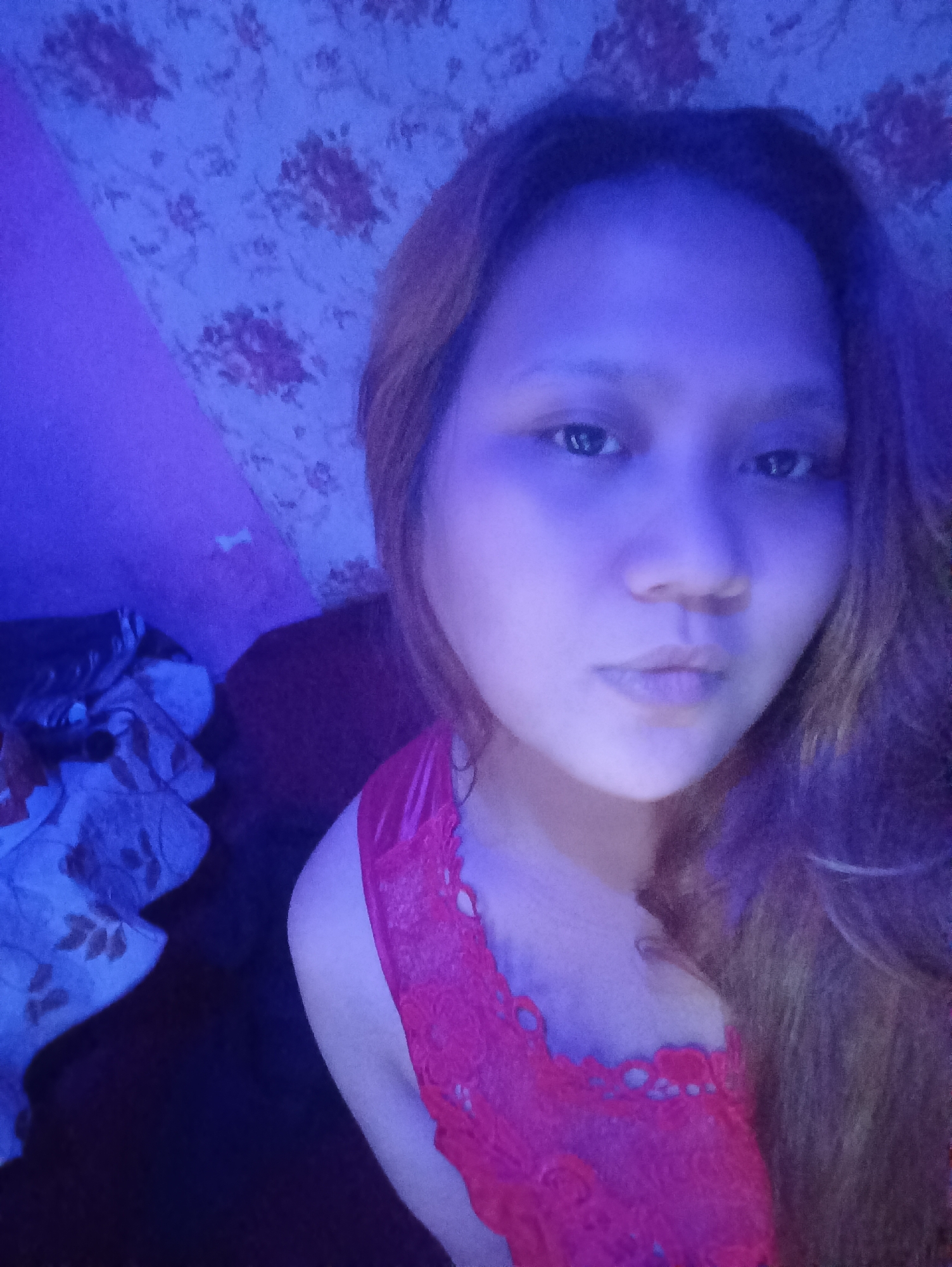 The image is a close-up, selfie-style photograph of a young woman. She has light brown hair and is wearing a red lace top. The lighting is dim and appears to be from a blue light source, casting a purplish hue over the scene. The background is indistinct, but suggests an indoor setting with patterned wallpaper and what appears to be some clothing or fabric to the left. There are no identifiable landmarks or elements that suggest a specific location within Depok, Indonesia. The woman's expression is neutral, and she is looking directly at the camera.