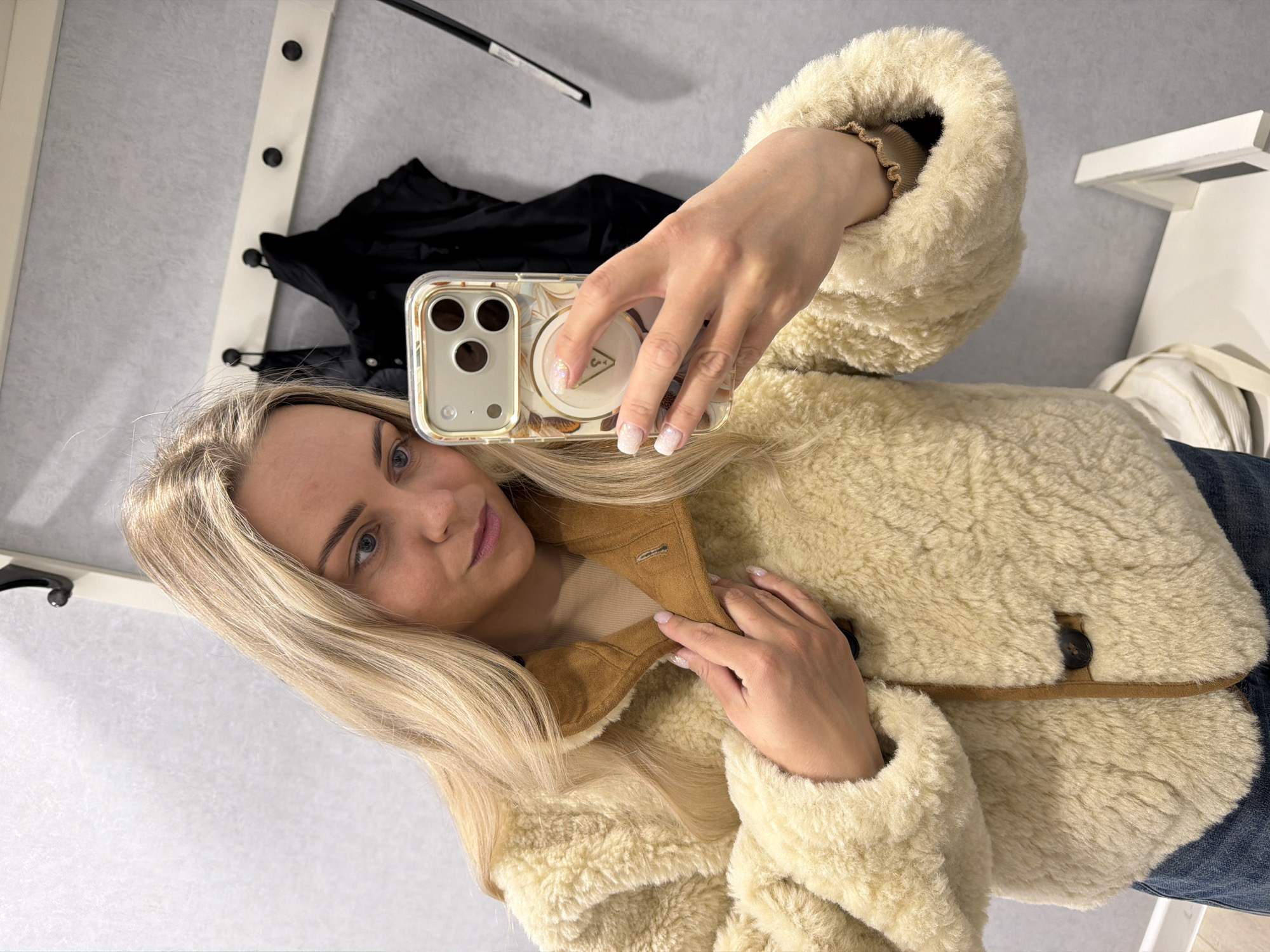 A fair-skinned woman with long blonde hair is taking a selfie indoors. She is wearing a cream-colored, sheepskin-like jacket with brown trim and buttons, and a light brown top underneath. She is holding a smartphone in a clear case with a floral pattern, covering most of her face. The phone has a circular magnet. She has light makeup and manicured fingernails.
The background includes a light gray wall, a white rack with black hooks, and a black garment hanging from it. There is also a white object with a flat surface and a white tote bag in the periphery.
The photograph appears to be taken inside what may be a dressing room or a retail space. There is no information in the image to indicate its location is in Votkinsk, Russia.