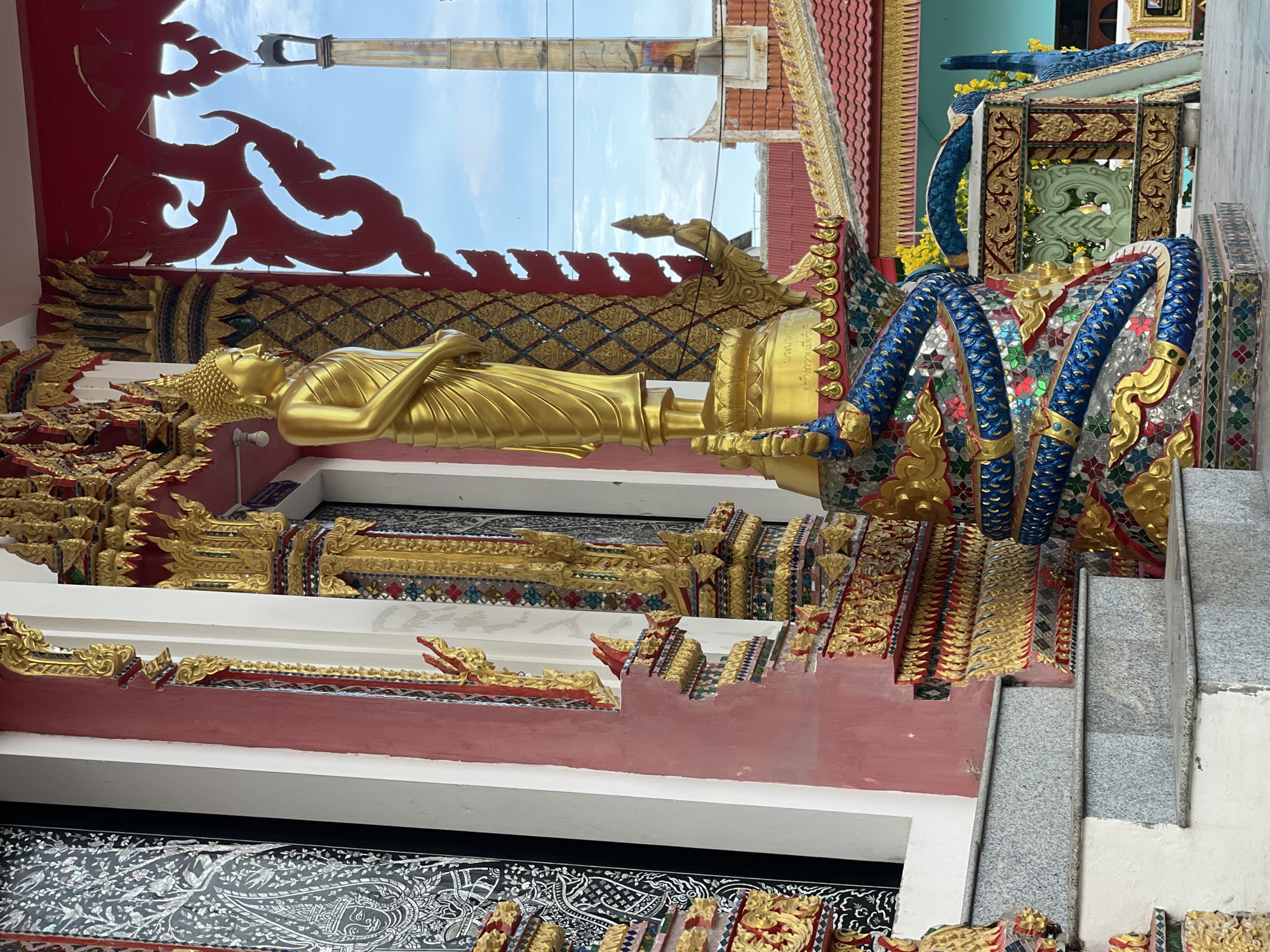 This image shows a statue of a golden Buddha in front of a temple in Pattaya City, Thailand. The Buddha is standing on a pedestal that is decorated with a blue and gold serpent. The temple is ornate with gold and red decorations. In the background, there is a tall structure that resembles a crematorium tower, with a red-tiled roof building beside it. The sky is blue with some clouds. The image is taken from a low angle, looking up at the statue and the temple.