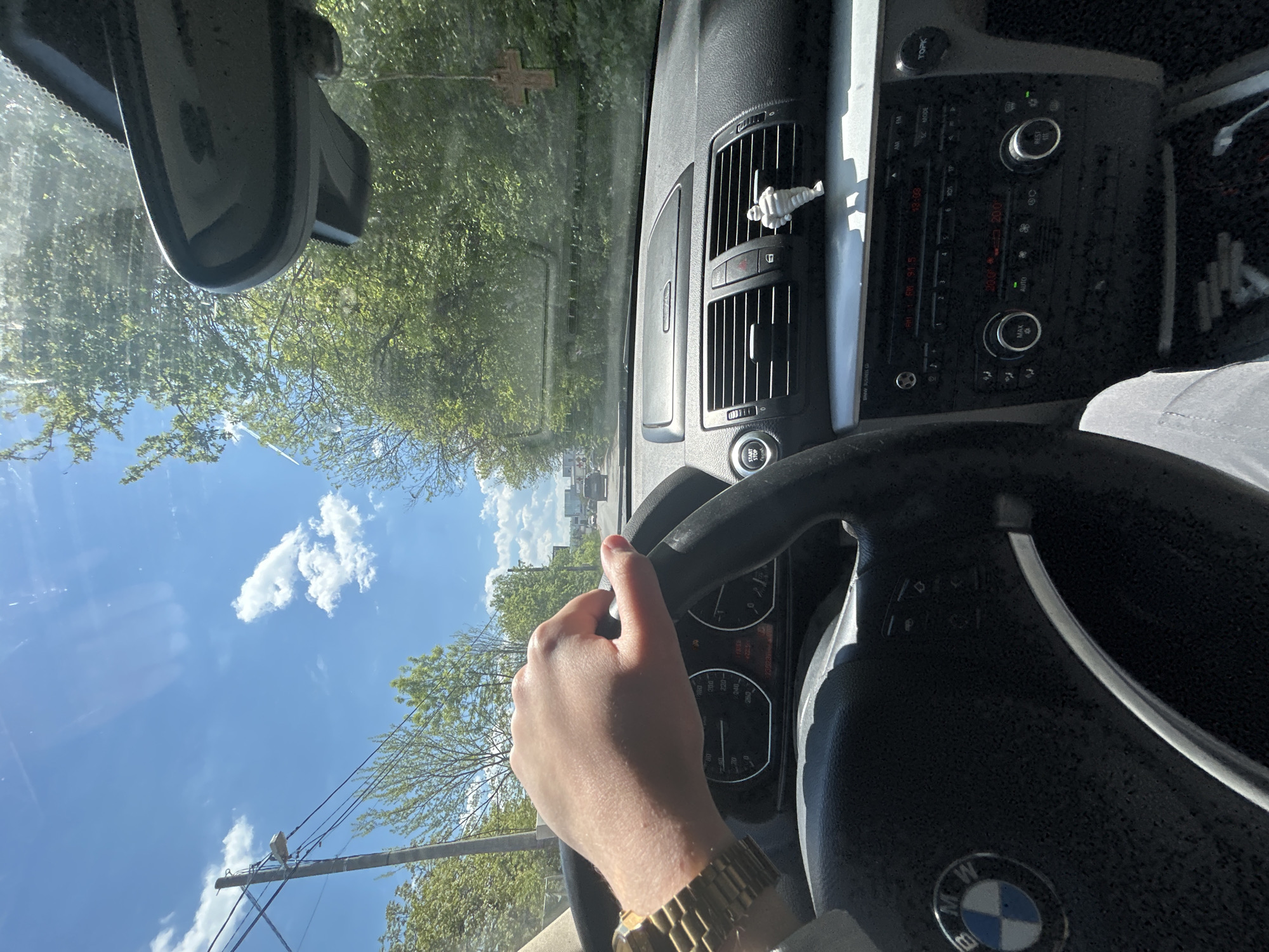 The image shows a view from inside a BMW car while driving. The driver's hand, wearing a gold watch, is visible on the steering wheel. The car's dashboard, including the air vents and radio controls, is in view. An astronaut figure is attached to one of the air vents. Through the windshield, trees and a blue sky with clouds are visible. A cross is hanging from the rear-view mirror. Based on the car model and the appearance of the surroundings, the location could be Sofia, Bulgaria. The car seems to be in motion on a road with trees and other buildings in the background.