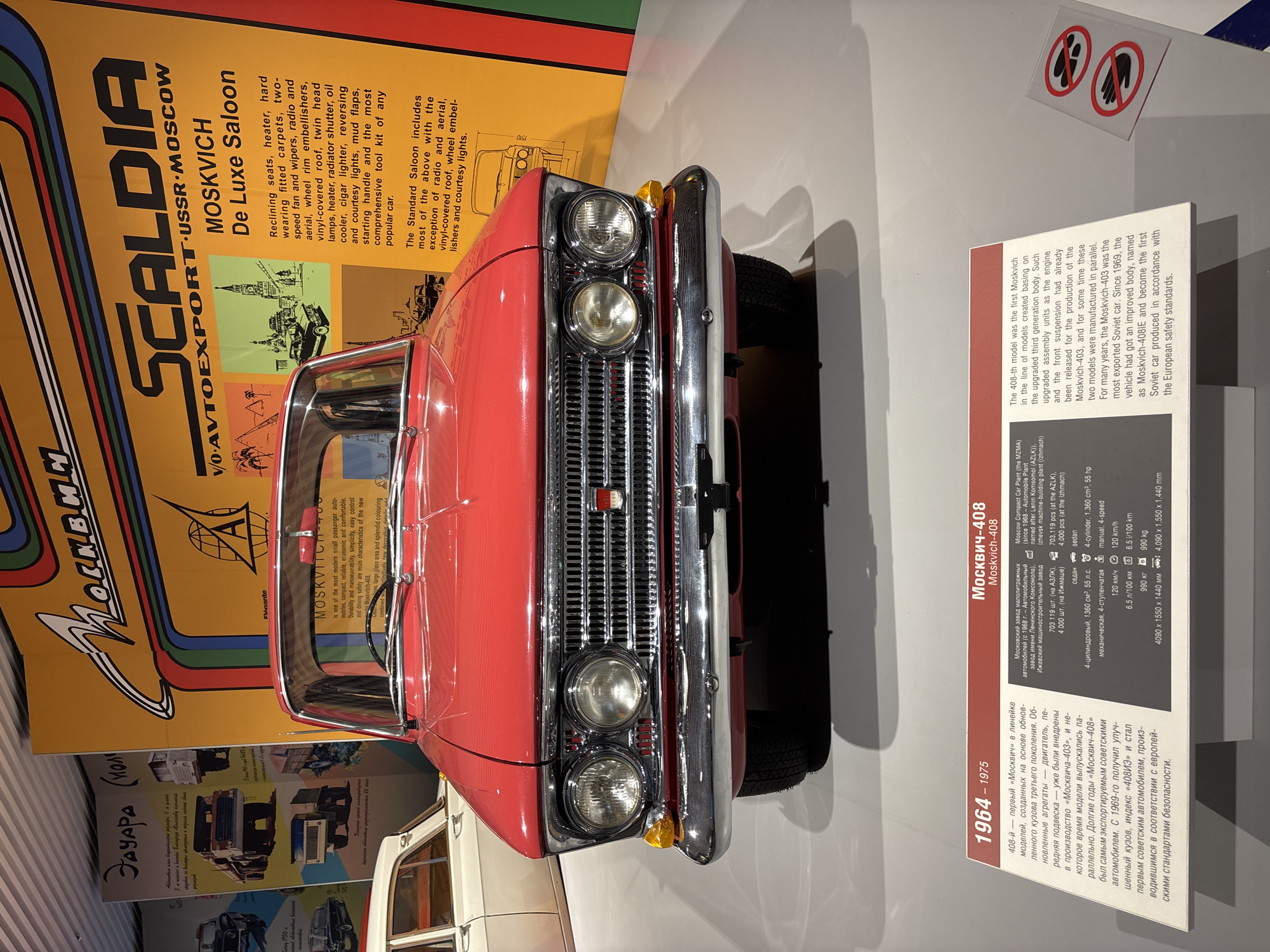This image shows a red Moskvich-408 car on display in a museum. The car is in the foreground, and behind it is a large yellow poster with text and images related to the car. The poster is in Russian and English and features the text "SCALDIA USSR MOSCOW MOSKVICH De Luxe Saloon". There is also a smaller white information plaque in front of the car, detailing its specifications and history. The museum is likely the Museum of Military Technology UMMC, located in Verkhnyaya Pyshma, Russia, which houses a significant collection of Soviet-era vehicles. The environment appears to be a well-lit exhibition hall with a white floor. There are no people visible in the image.