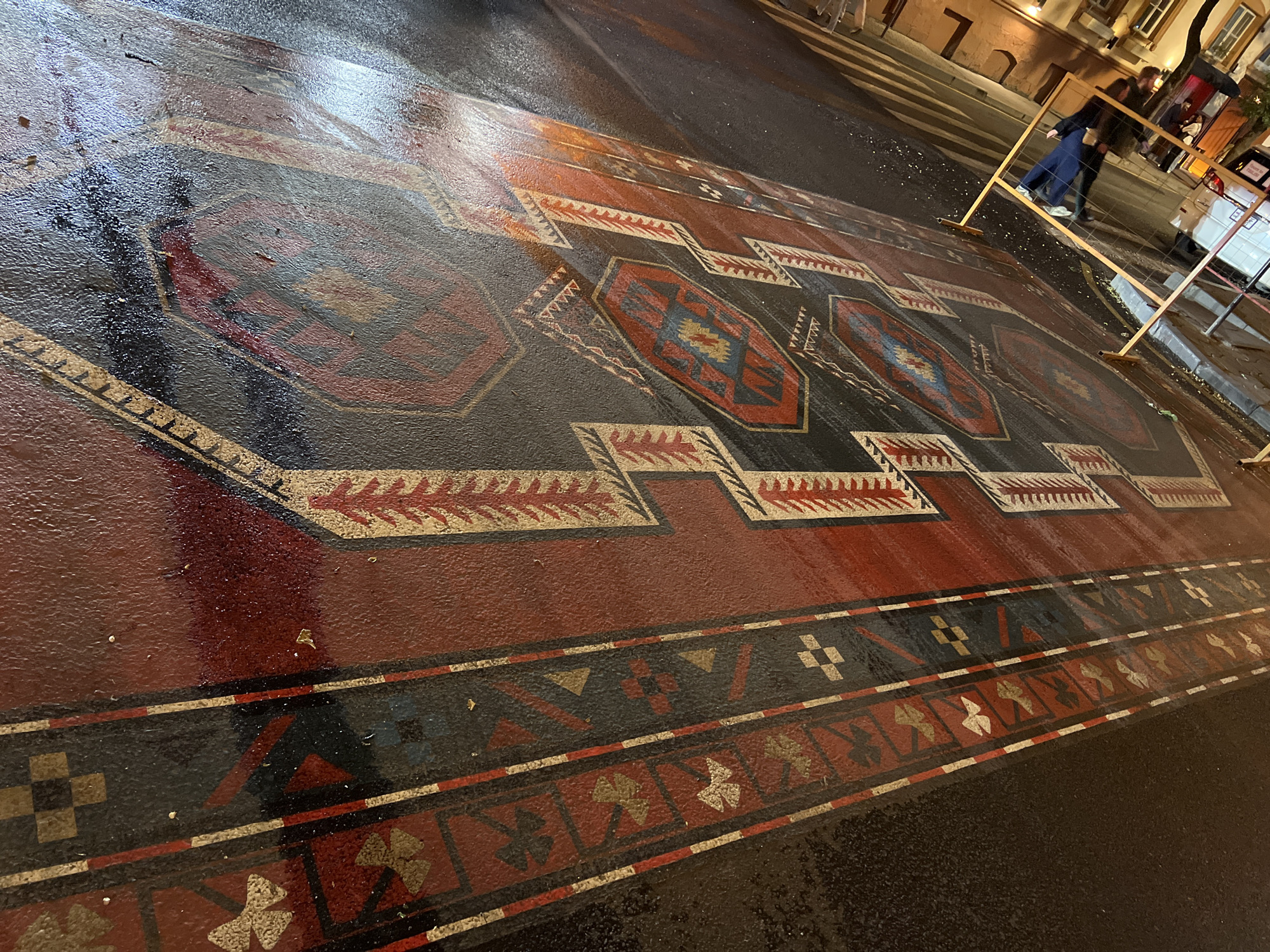 The image shows a large, colorful carpet painted directly onto a wet asphalt street. The design resembles a traditional Armenian rug, featuring geometric patterns and motifs in red, black, and other colors. The street surface is reflective due to the moisture, enhancing the visual impact of the artwork. In the background, a street scene unfolds with pedestrians walking near a barrier marking the edge of the carpet art, suggesting the artwork is on display for public viewing in a sectioned-off area. The context of the scene, the city of Yerevan, Armenia, is known for artistic displays in public spaces. The street architecture and evening lighting further suggest an urban environment.