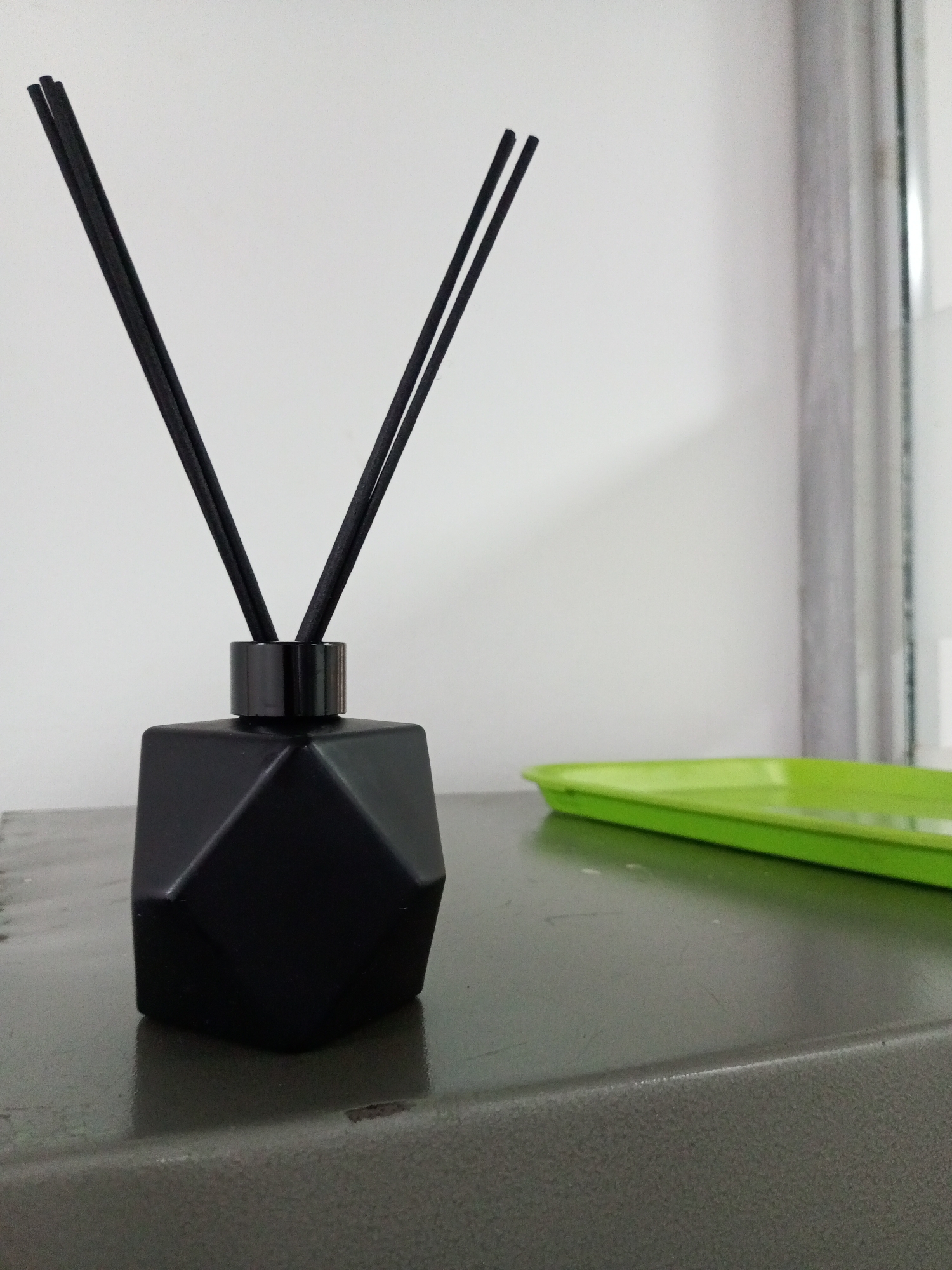 In Bukuru, Nigeria, a black, faceted diffuser bottle with four black reeds stands on a grey surface. To the right of the diffuser, a bright green plastic tray is visible. The background consists of a plain white wall.