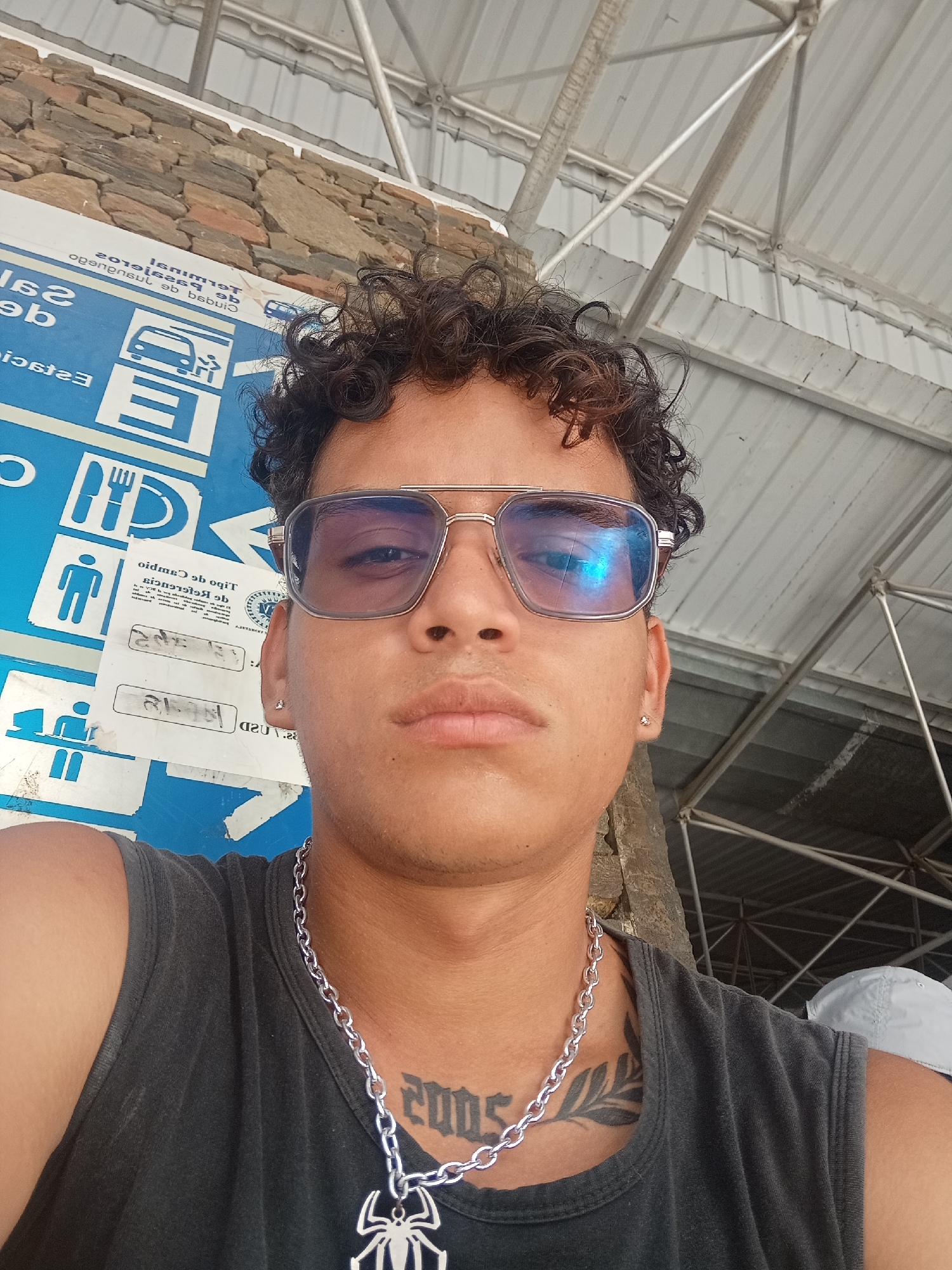 Here is a description of the image:
A selfie perspective shows a young man wearing tinted aviator glasses, a silver chain necklace with a spider pendant, and a dark tank top. He has short, curly dark hair and small earrings. The background includes a sign with bus and dining symbols, partially obscured text, and a stone wall. A metal framework and roof are also visible above. The setting appears to be in the city of Juan Griego, Venezuela, evidenced by the text on the sign "Terminal de pasajeros de bebus" (Passenger terminal of bebus, a bus company in Venezuela) and the location identification. The subject seems to be posing for the photo.