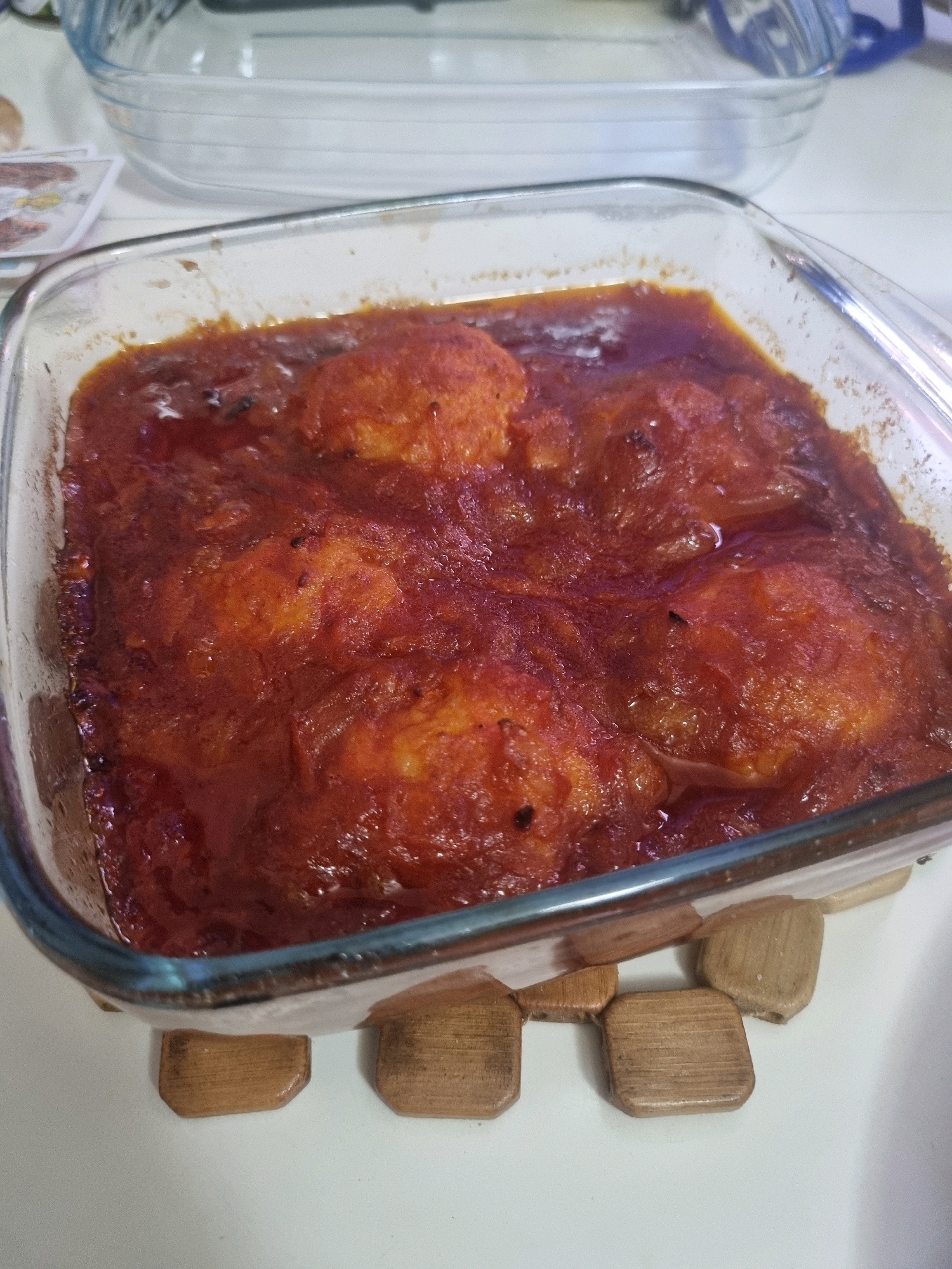 A close-up, overhead shot shows a square glass baking dish filled with a saucy dish containing several large, round, golden-brown food items. The dish is placed on a white surface, supported by four small, wooden blocks. The food appears to be stewed or baked, with a thick, red sauce coating the entire contents. The items within the dish are submerged in the sauce and their texture suggests they might be dumplings, meatballs, or some form of bread or dough. The background shows another, larger glass baking dish, partially visible and empty. To the left of the main dish, some papers with images are also visible. The lighting is bright, suggesting an indoor setting, likely a kitchen. No people or specific location details are discernable within the image itself.