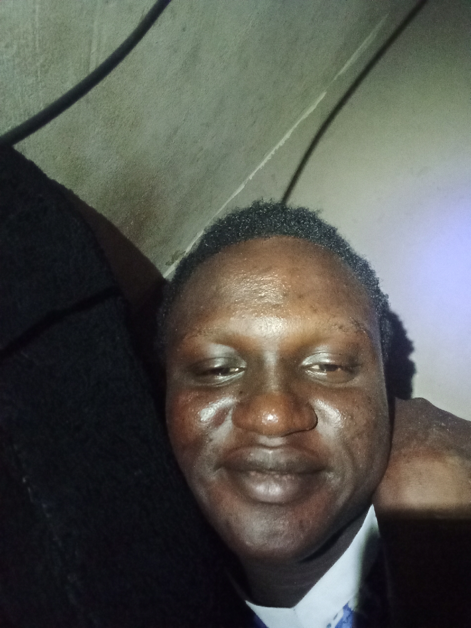 A close-up selfie of a dark-skinned man with short, dark hair. The man's face is well-lit, showing some skin texture. The background is blurry. Part of a dark fabric is visible on the left side of the image, and a light-colored wall or ceiling is in the background.
Location: Unknown, Unknown.