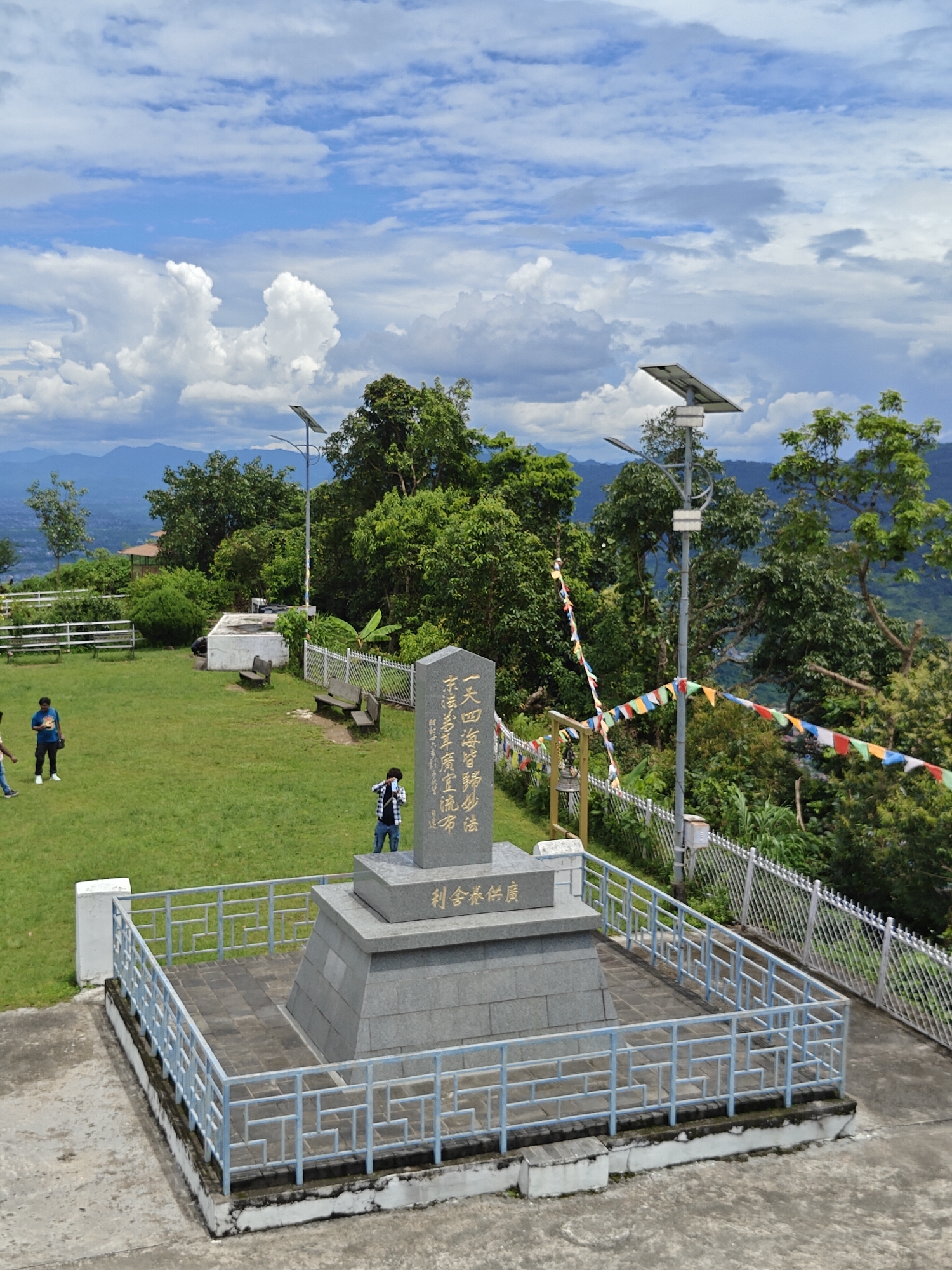 The image shows a monument in Pokhara, Nepal. The monument is made of granite and has inscriptions in Chinese. It is surrounded by a blue metal fence. There is a grassy area with benches and trees around the monument. In the background, there are mountains and a cloudy sky. Three individuals are visible in the image: two men standing on the grass and one man taking a picture of the monument. The man taking the picture is wearing a plaid shirt and jeans. One of the men on the grass is wearing a blue t-shirt and black shorts, while the other is wearing a light-colored shirt and black pants. Solar-powered streetlights are visible on poles throughout the area. Prayer flags are strung across a fence in the middle ground.