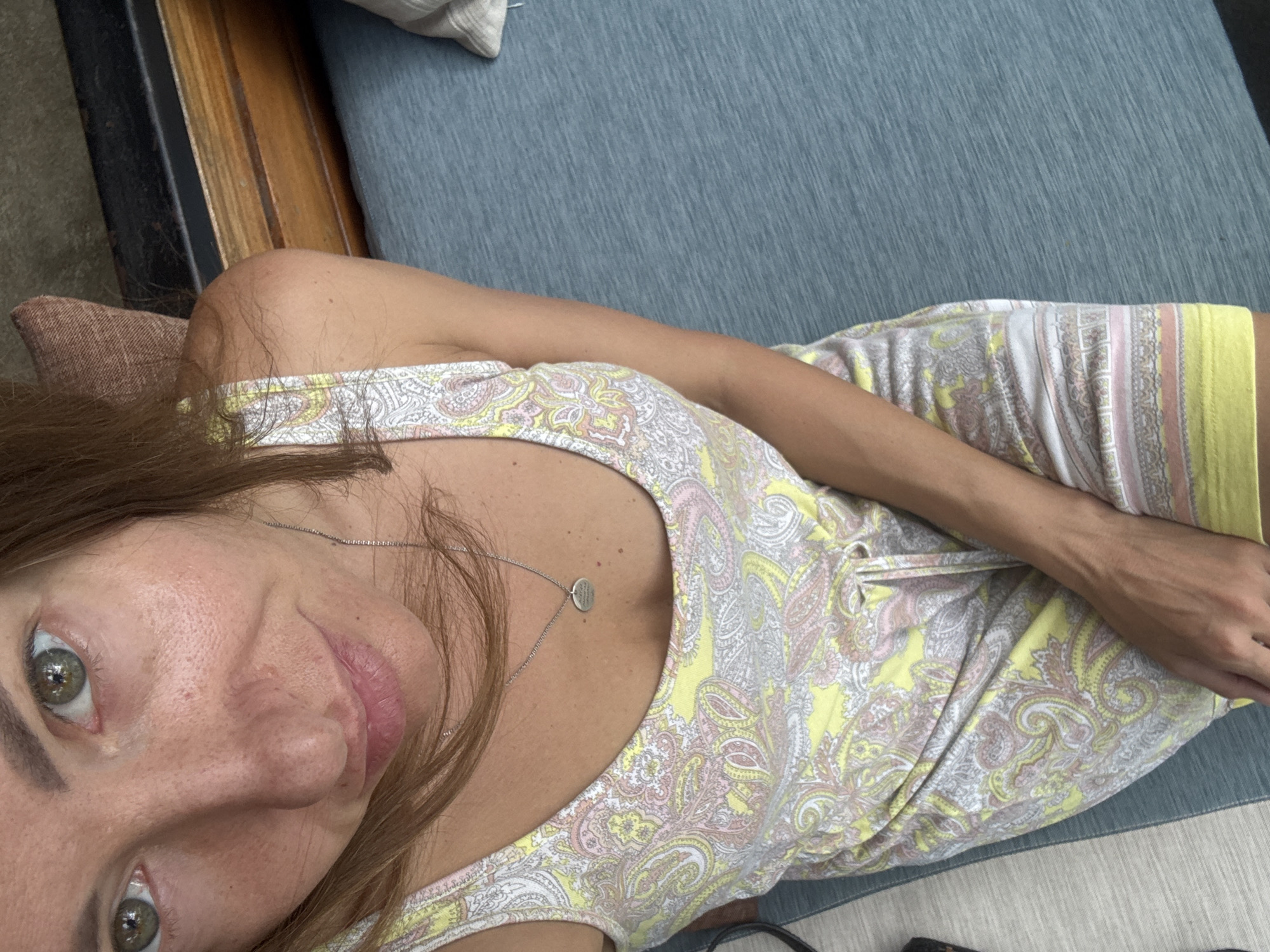 The image is a first-person perspective shot of a woman wearing a light yellow and pink paisley-patterned dress. She is seated on a blue cushion with a wooden frame. The environment suggests an indoor or sheltered outdoor setting. The image was taken in North Kuta, Indonesia. The woman is wearing a necklace.