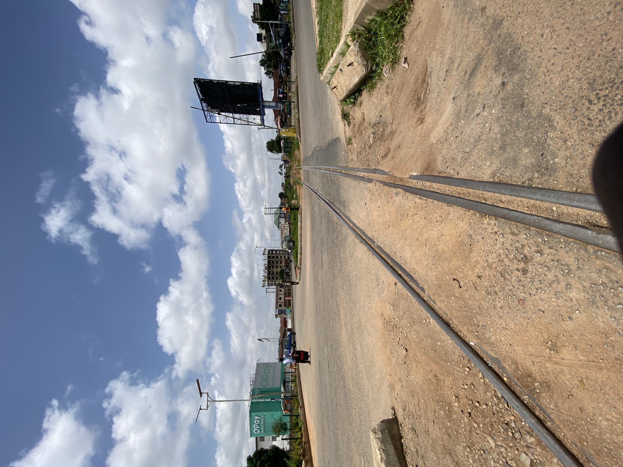 A vertical, eye-level, full shot captures a dirt road with train tracks running parallel to the viewer, leading toward a distant intersection under a bright, cloudy sky. In the foreground, the train tracks are set in dusty, reddish-brown earth. The road surface transitions to asphalt as it approaches the intersection.
To the left of the intersection, a large, bright green billboard with "Opay" prominently displayed is visible. A lamppost with solar panels stands near this billboard. Across the road, to the right, a large, dark billboard stands on a tall pole, its surface appearing to be in disrepair.
Several vehicles are scattered around the intersection. A blue car is stopped on the road, and a motorcycle with a rider is visible near the "Opay" billboard. Further into the distance, buildings of various sizes and stages of construction are present. Some appear to be residential or commercial structures, while others are under development, with scaffolding visible.
The environment suggests a developed area within a city, likely experiencing active construction or undergoing development. The presence of both a road and train tracks indicates a transportation hub or an area with a history of rail activity. The overall scene depicts a typical urban landscape with elements of commerce, infrastructure, and ongoing progress. The image was taken in Jos, Nigeria.