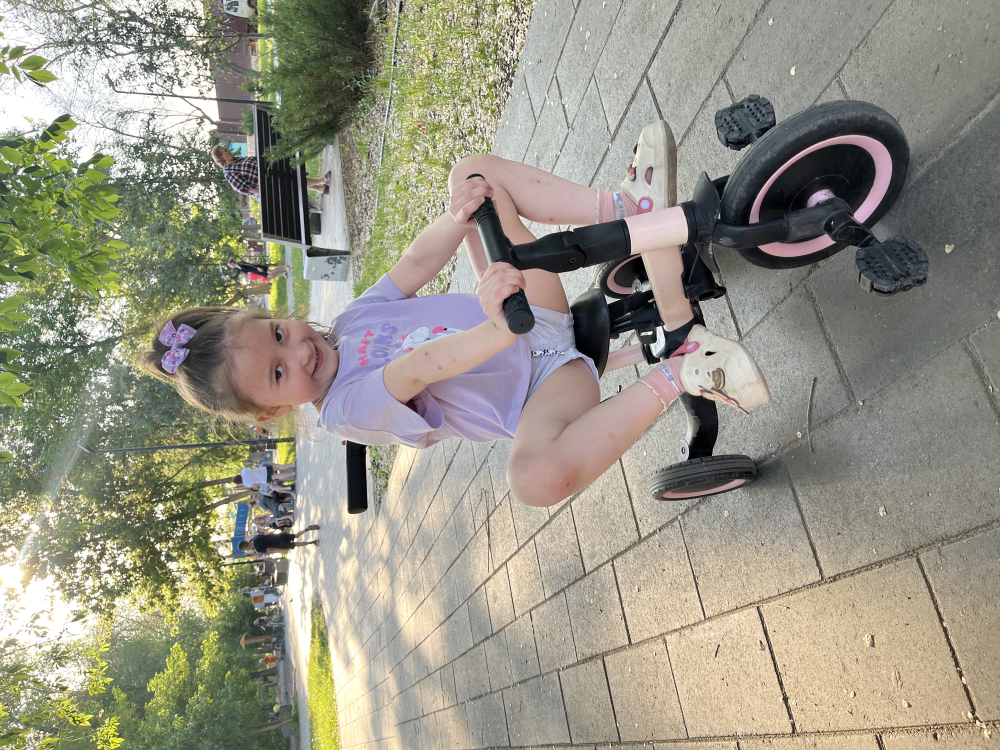 A young girl is sitting on a tricycle in what appears to be a park or public walkway. The tricycle is black and pink, with the girl facing the camera and holding the handlebars. She is wearing a light purple t-shirt with a cartoon graphic, grey shorts, and white shoes with pink socks. Her hair is pulled back with a patterned bow.
The ground beneath her is a paved walkway made of grey stone blocks. In the background, there are trees and other people, including a person sitting on a bench. The lighting suggests it is daytime, possibly late afternoon or early evening.
Based on the identified location (Novokuybyshevsk, Russia), this scene is likely taking place in a public park within that city.