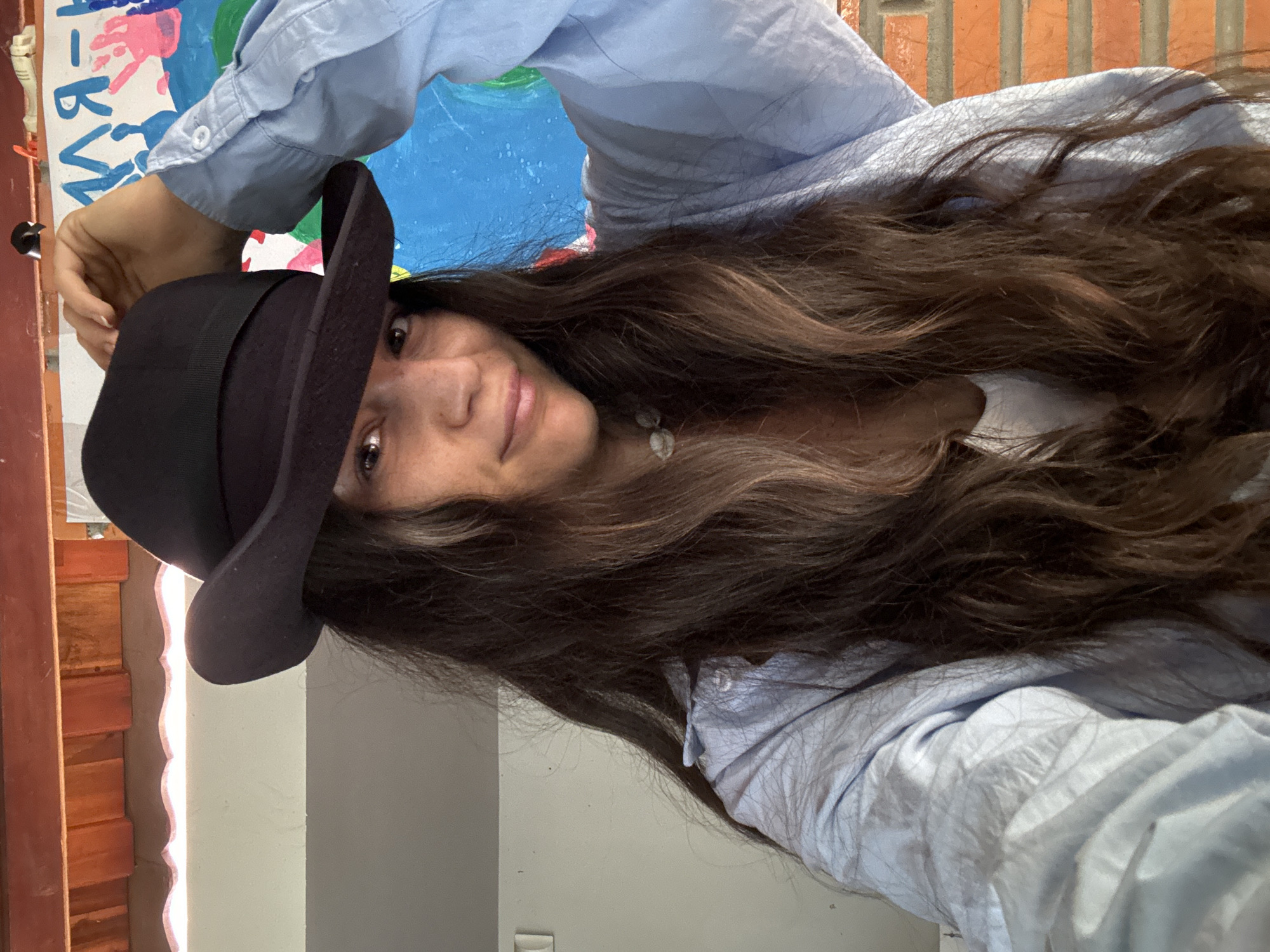 A woman is posing for a selfie outdoors, wearing a black fedora hat tilted to the side. She has long, dark brown hair and is wearing a light blue button-up shirt over a white top. She is looking at the camera with a slight smile. Her right hand is raised, touching the brim of her hat. In the background, there is a colorful mural or painting on a white canvas, and a brick wall. The scene appears to be on a covered patio or porch, with wooden beams visible overhead. The overall impression is casual and playful.