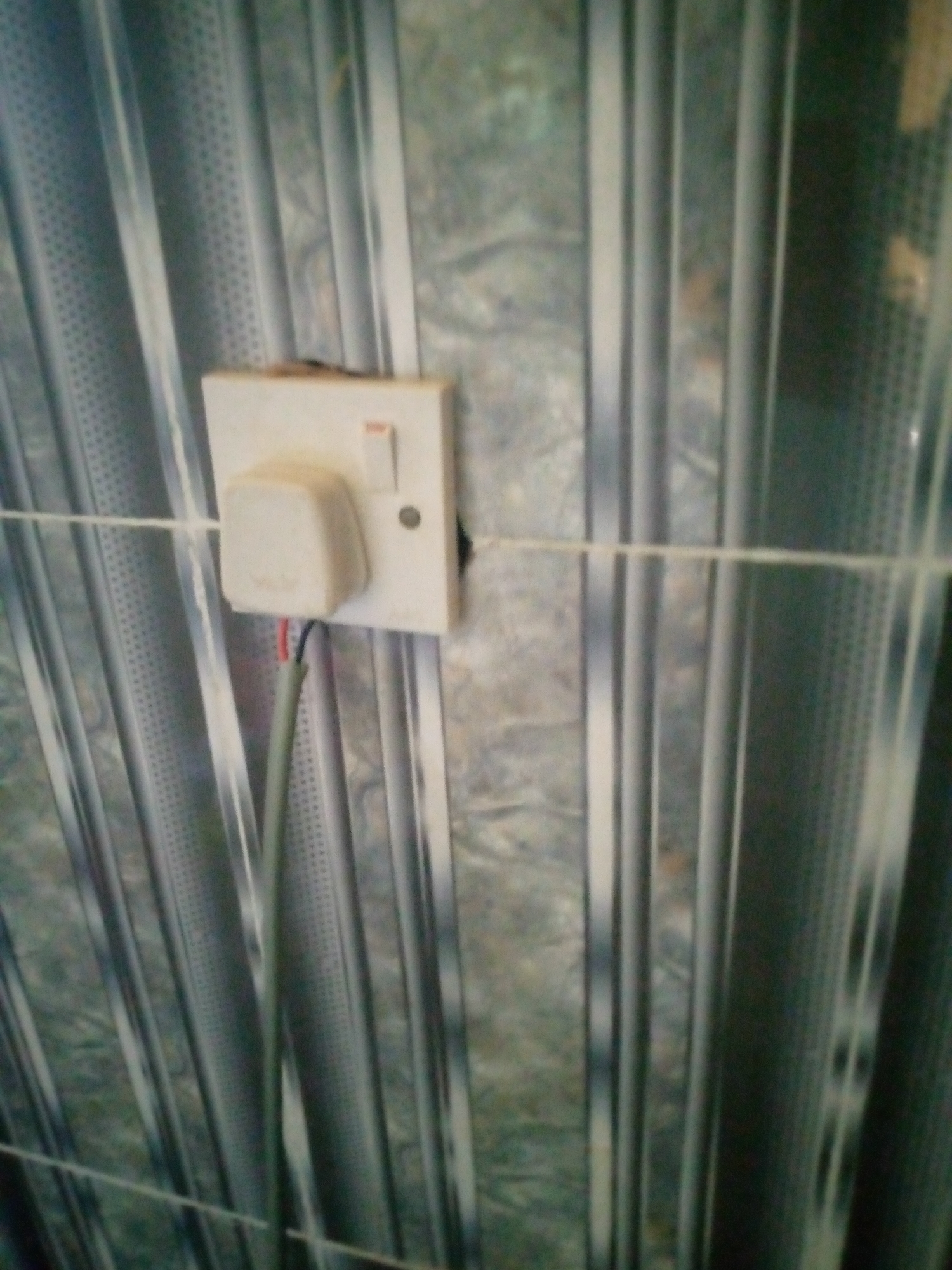 The image shows a close-up of a white electrical outlet with a plug inserted. The outlet is mounted on a wall that appears to be made of a metallic, corrugated material. A thin white string is stretched horizontally across the image, passing in front of the outlet. Wires are visible emerging from the plug and extending downwards. The lighting suggests an indoor environment. The location is identified as Jalingo, Nigeria.