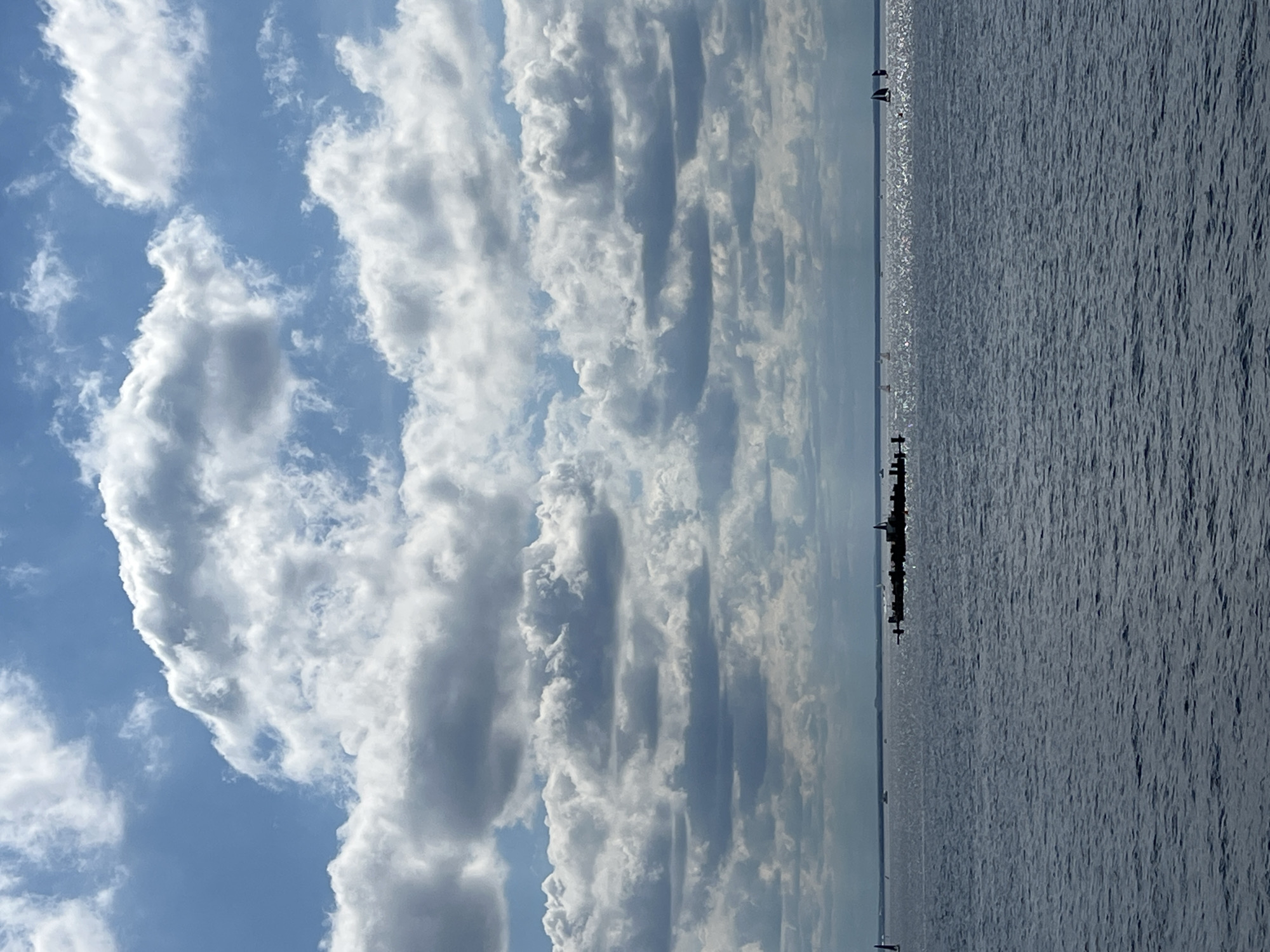 The image depicts a wide expanse of calm, deep blue water under a partly cloudy sky. Several sailboats are visible on the horizon, their white sails contrasting with the dark water. In the mid-ground, a long, dark structure, possibly a pier or breakwater, juts out into the water, with several smaller structures or boats clustered on it. The clouds are varied, with large, white, fluffy cumulus clouds dominating the upper portion of the sky, interspersed with darker, more diffused cloud formations. The lighting suggests it is daytime, likely with the sun partially obscured by clouds, creating a bright but not overly harsh illumination. The overall scene conveys a sense of tranquility and openness, characteristic of a coastal or large body of water. The location is unknown and the presence of any specific landmarks or identifiable city features is not discernible.